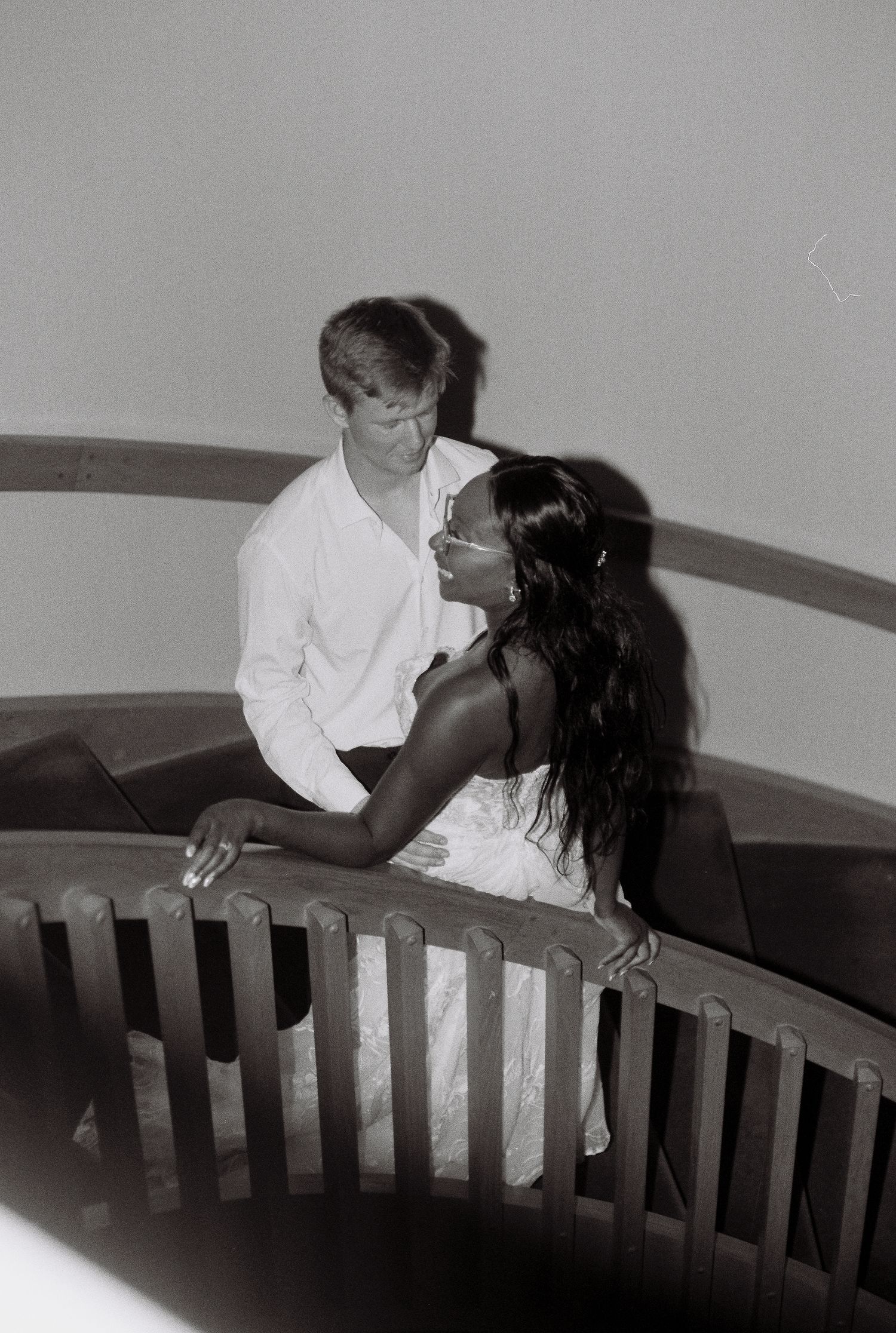 Black and white photo of an interracial couple sharing a romantic moment on a curved staircase railing.