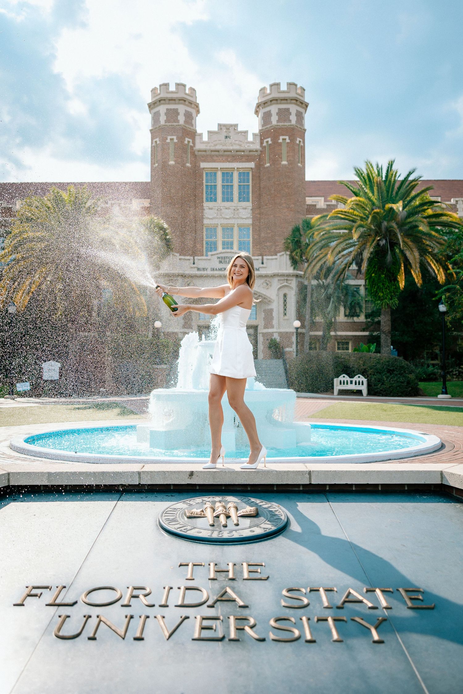 A fountain splashes in front of historic Florida State University buildings with palm trees and blue sky.