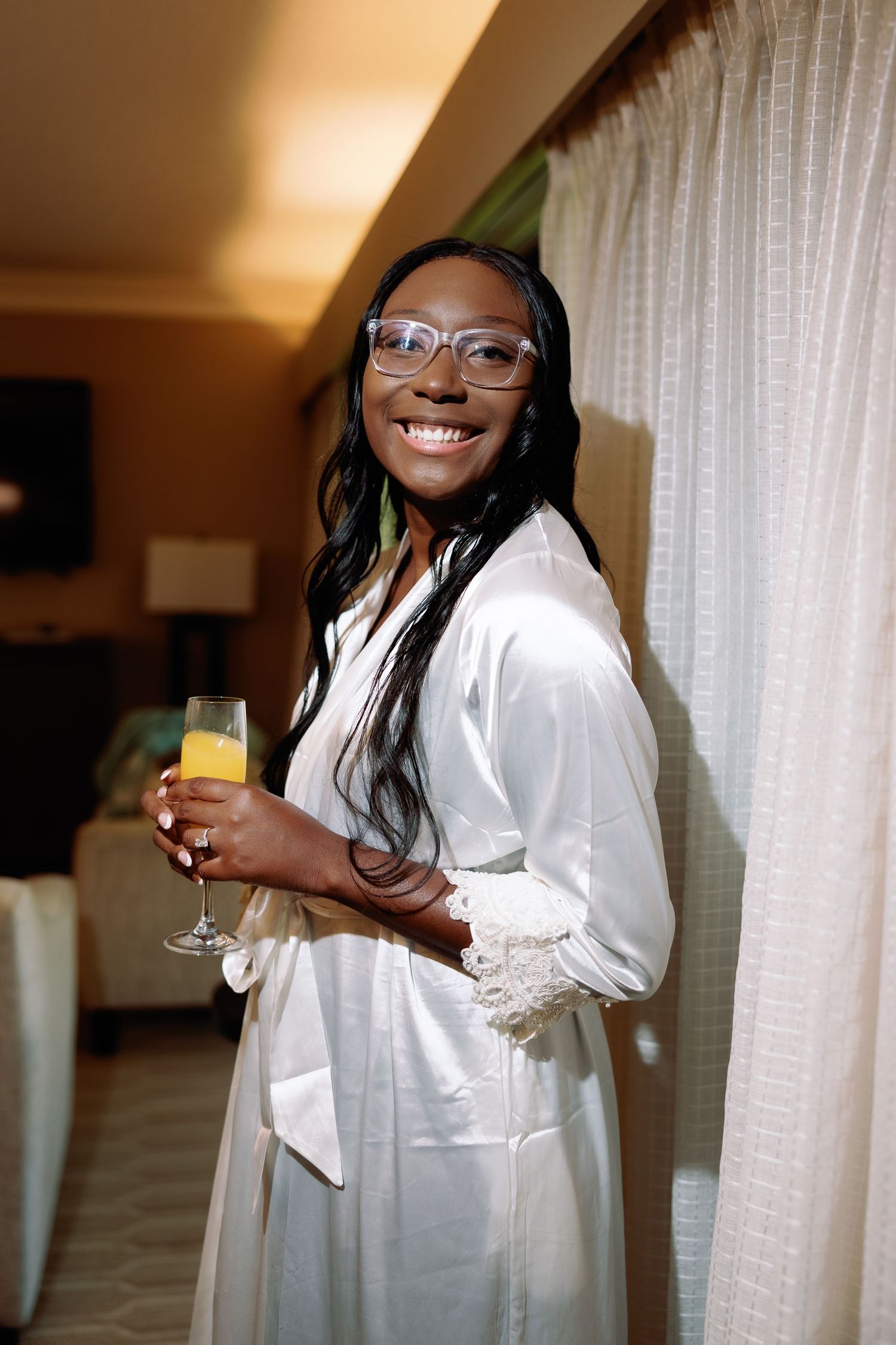 Radiant Black bride in white satin robe and glasses holds mimosa glass, smiling in hotel room.