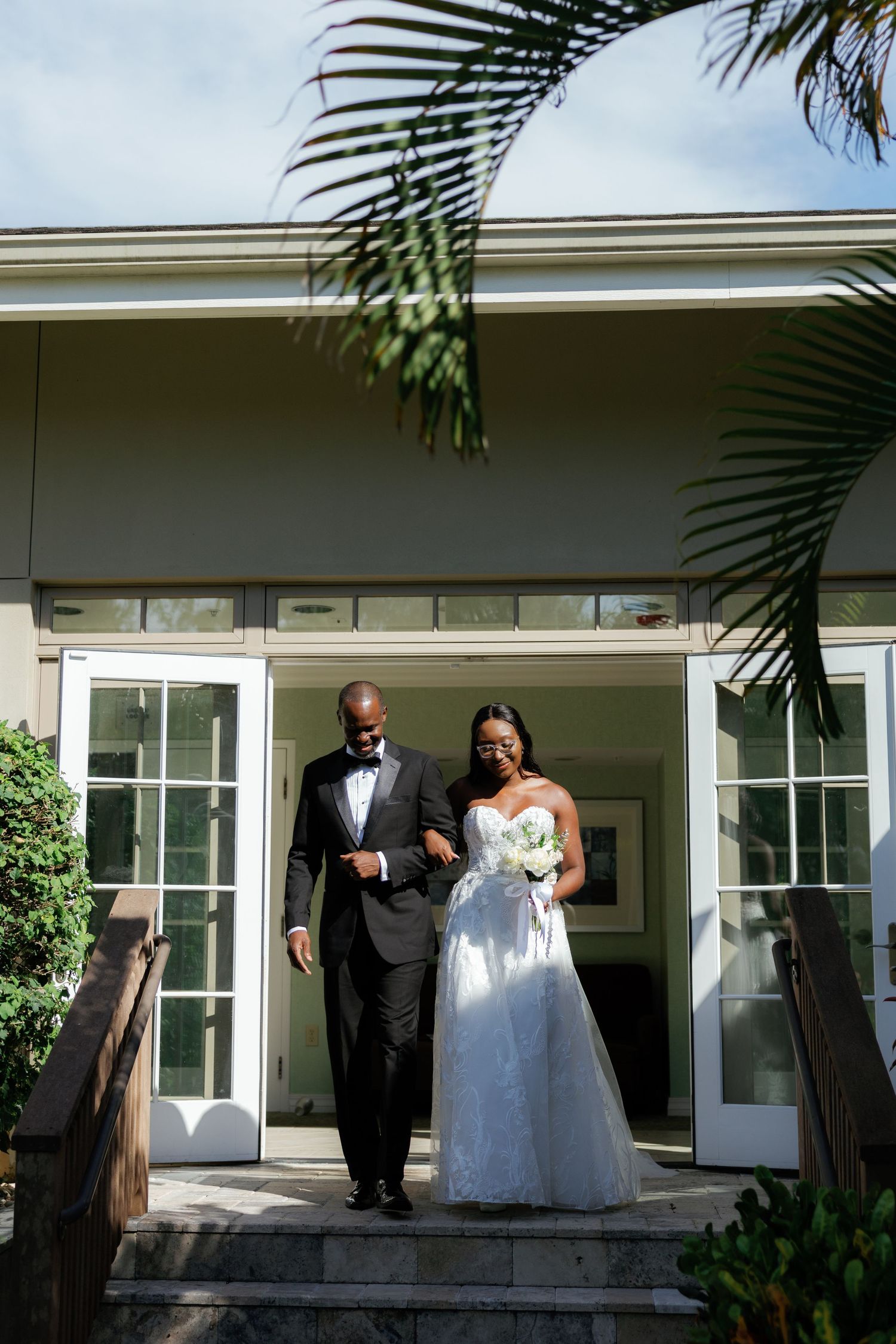 Bride and groom exit building in wedding attire, she holds white bouquet, palm trees frame elegant venue steps.