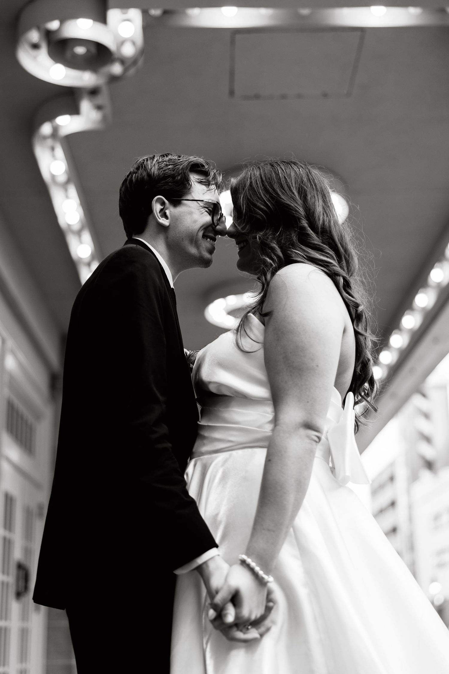 Black and white photo of smiling newlyweds holding hands and touching noses under marquee lights.
