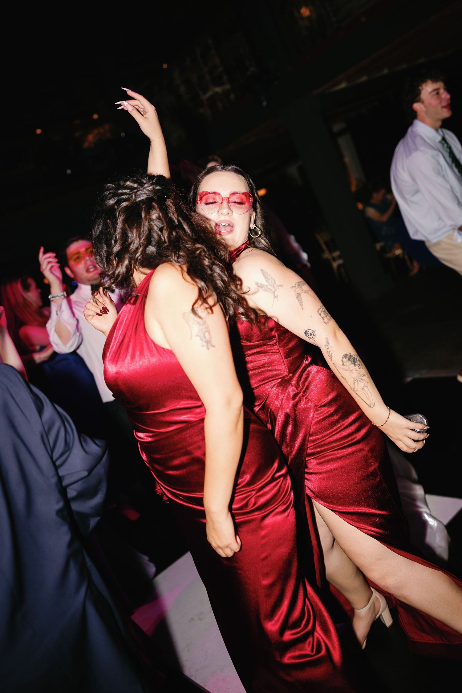 Two women in red satin dresses dancing energetically at a formal event, wearing matching red sunglasses.