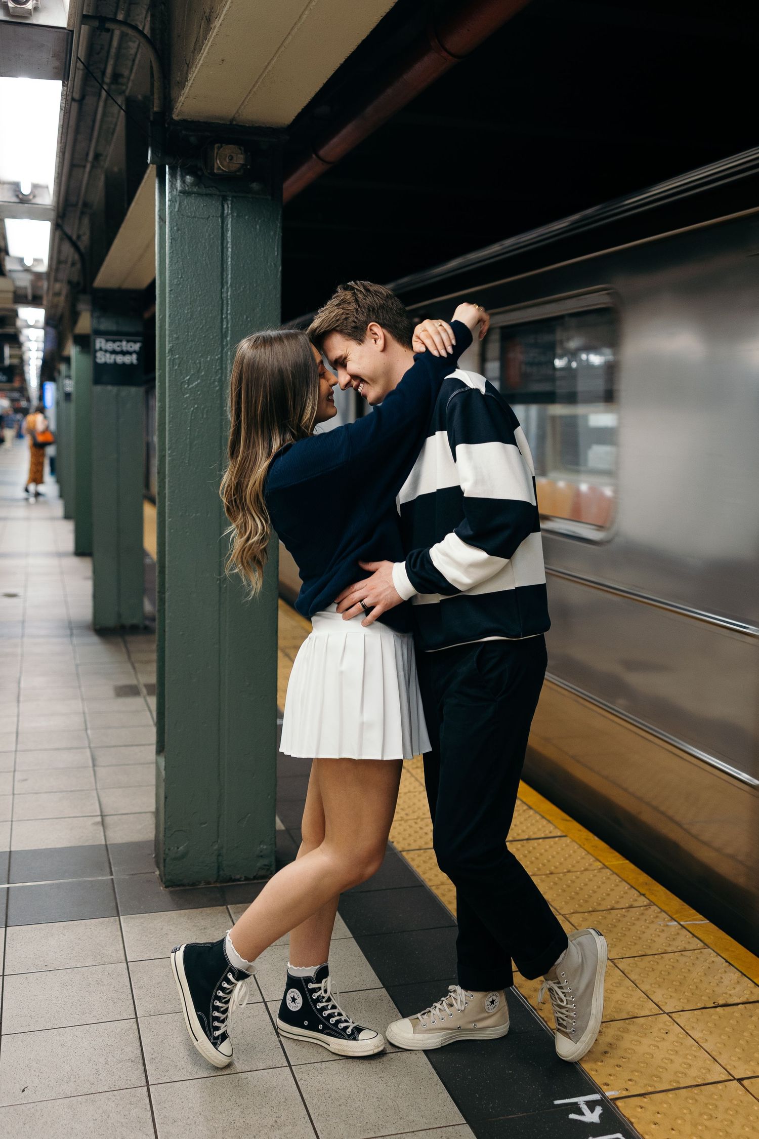 A couple in casual attire sharing a romantic moment on a subway platform with white tile walls.