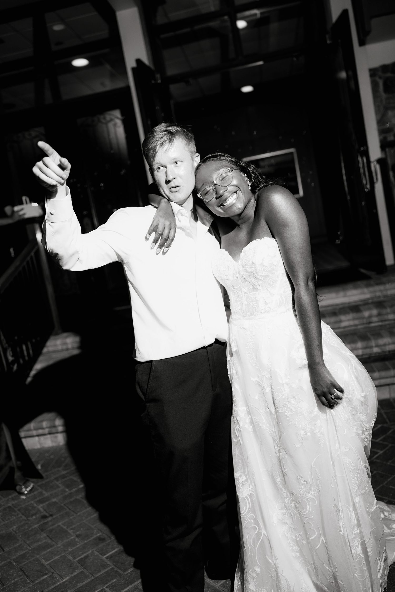 Black and white photo of a bride in lace gown and groom in white shirt posing joyfully at their wedding reception.