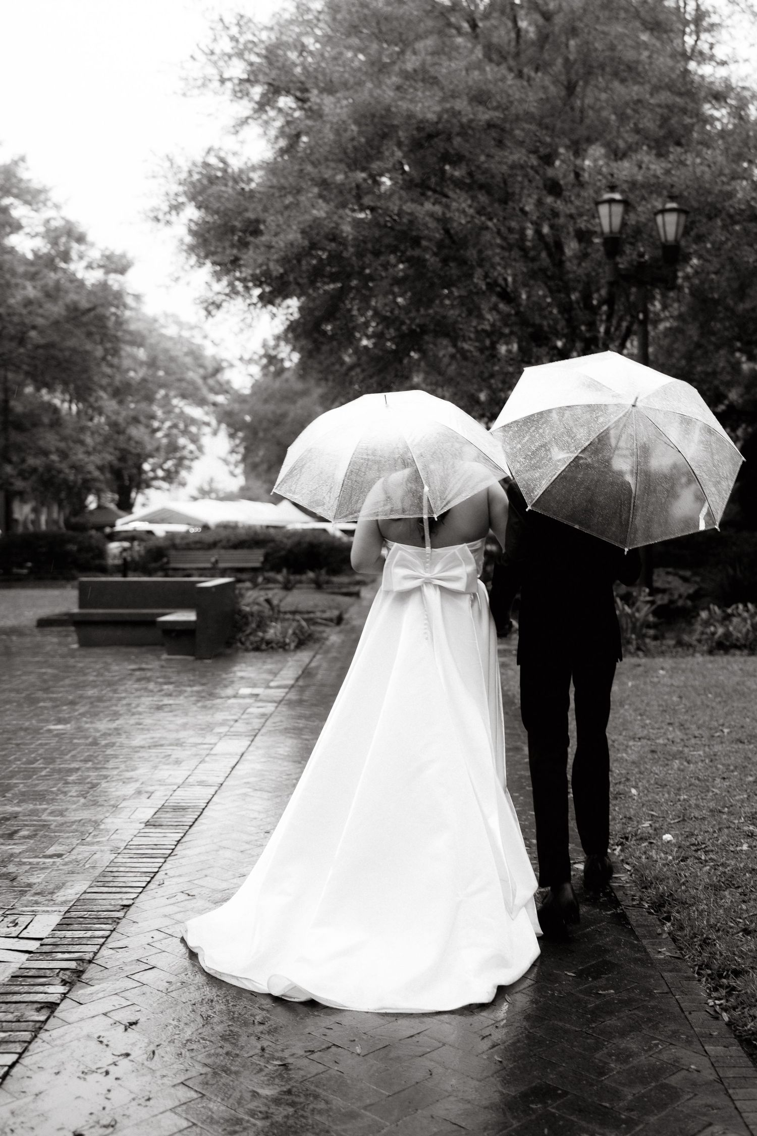 Black and white photo of bride and groom walking in rain holding clear umbrellas on a cobblestone path.