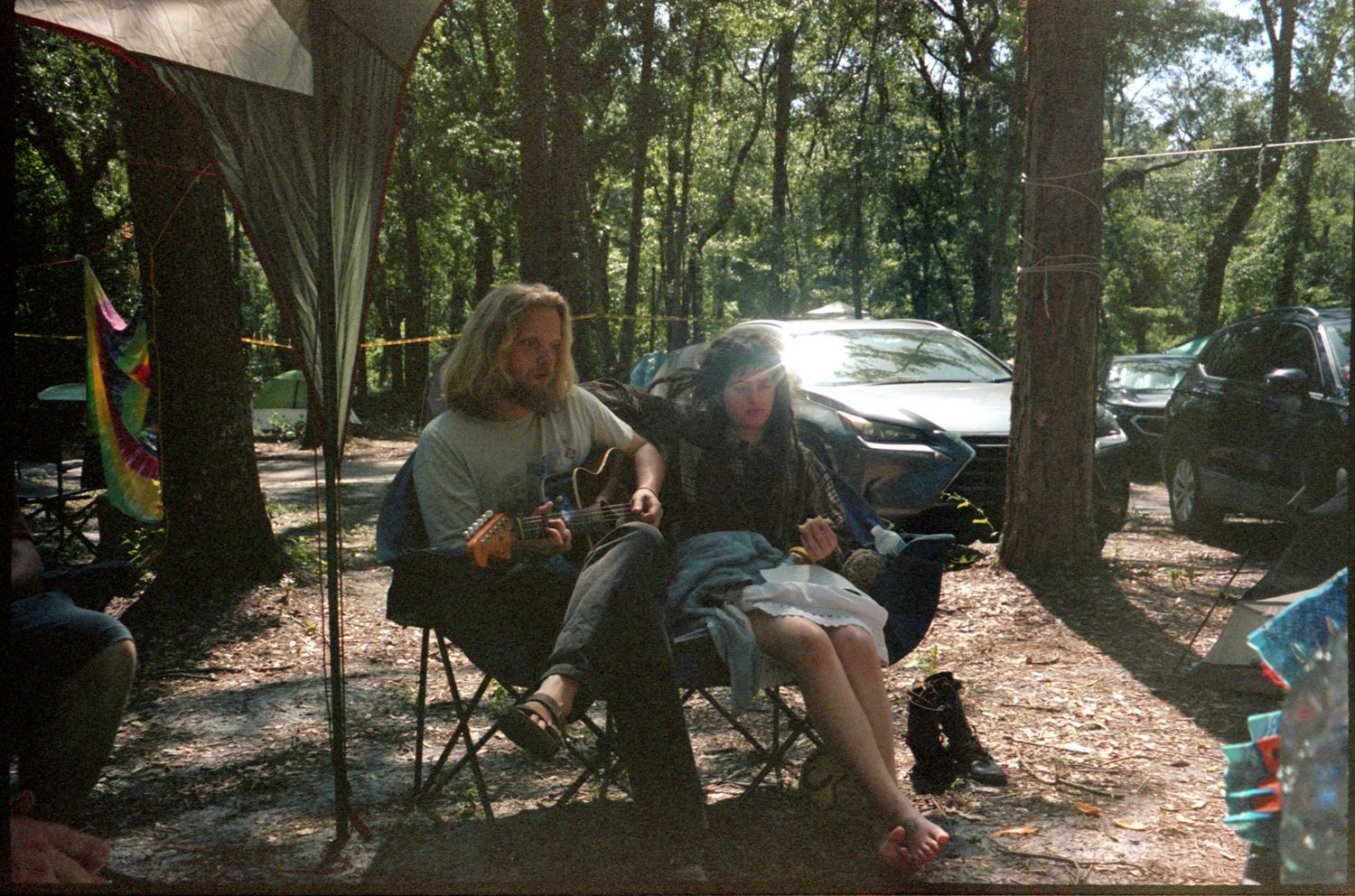 People sitting in camping chairs among trees at an outdoor gathering with cars parked in the background.