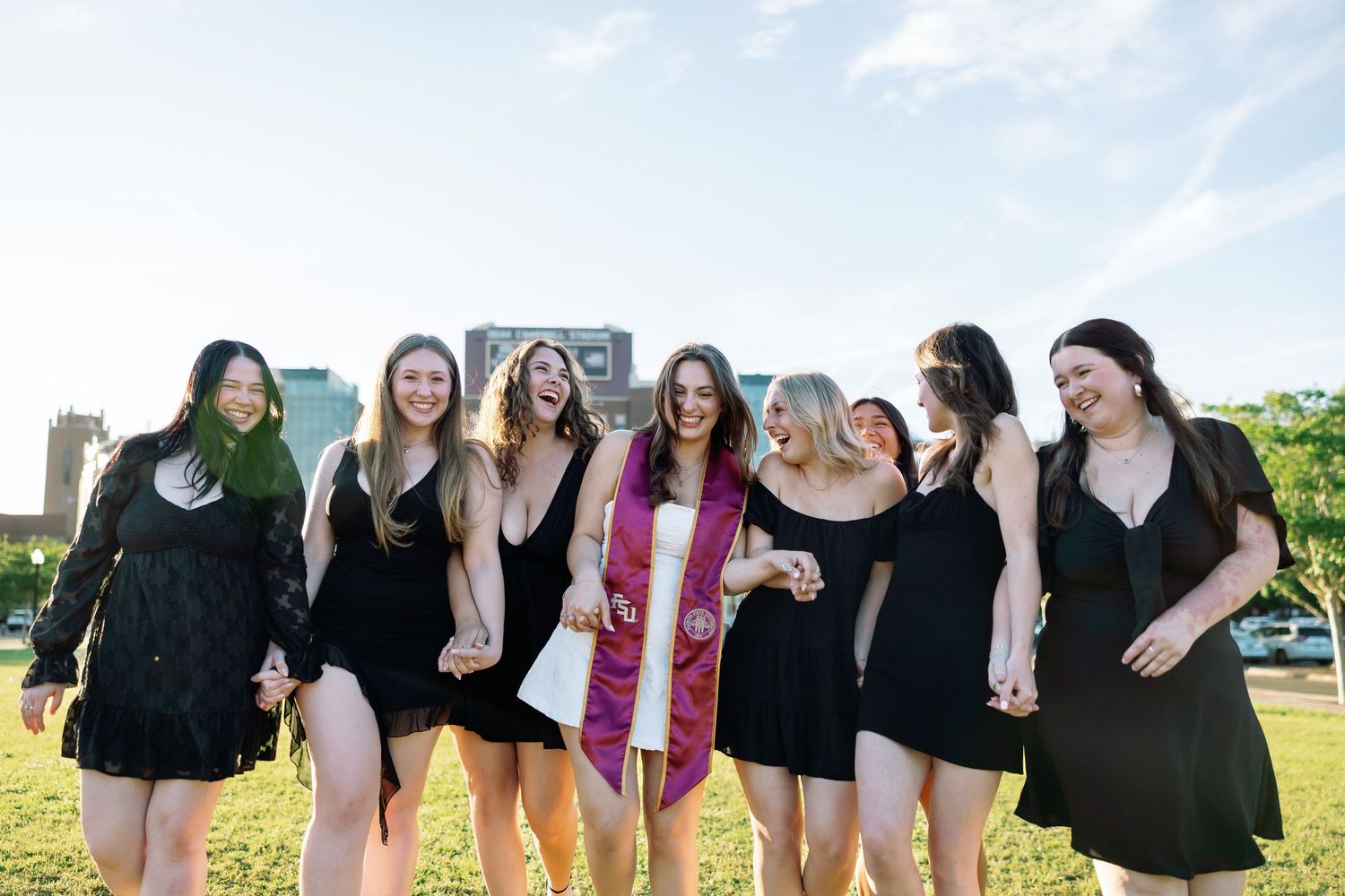 Group of seven women in black dresses celebrating graduation outdoors, one wearing white sash with purple stole.