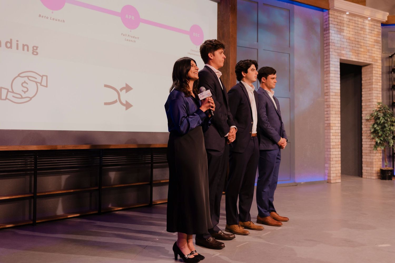 Professional speakers and panel participants stand at a business presentation in a modern corporate event space.