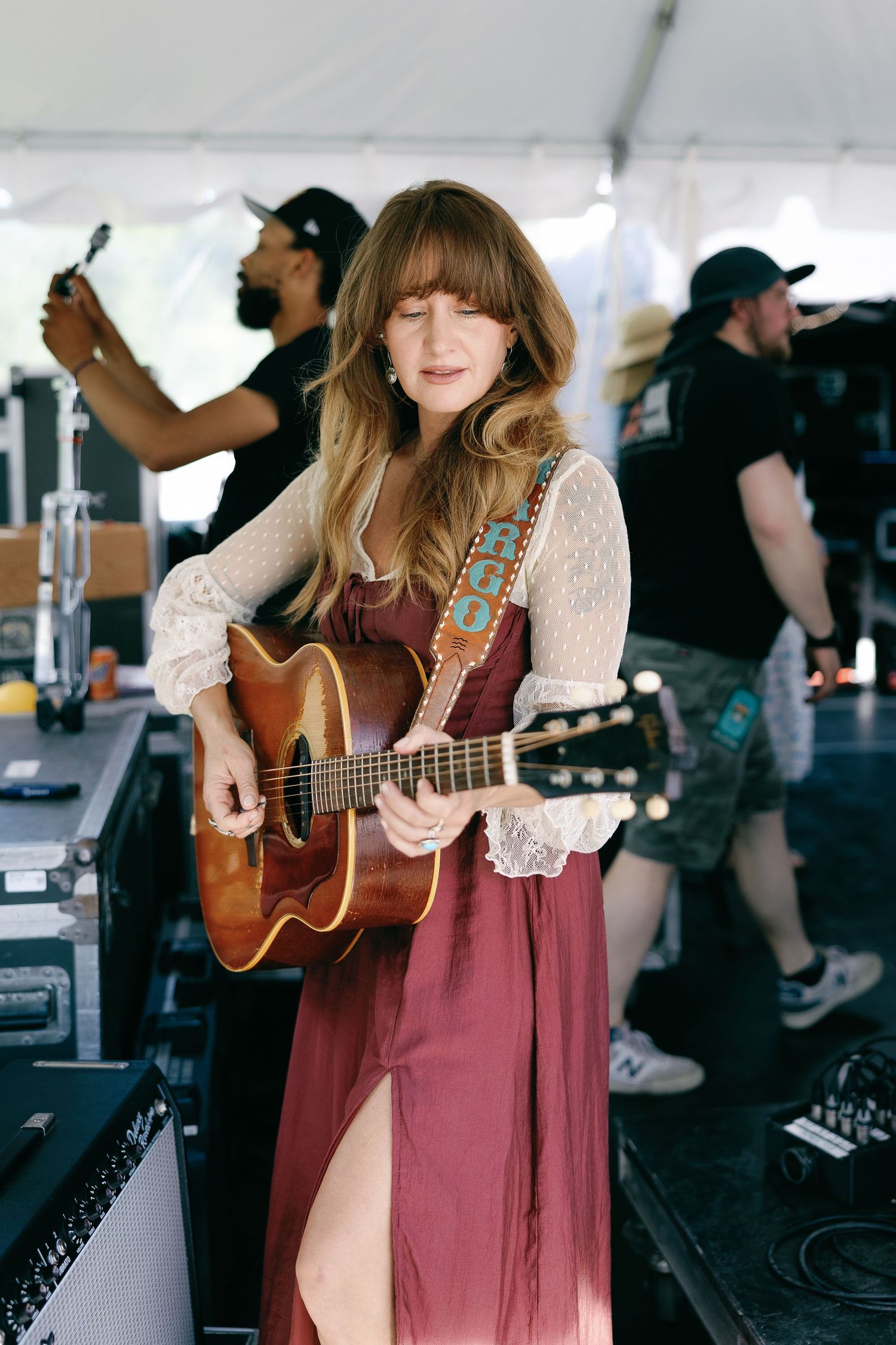 A woman in a burgundy dress plays acoustic guitar under a tent at an outdoor music festival.
