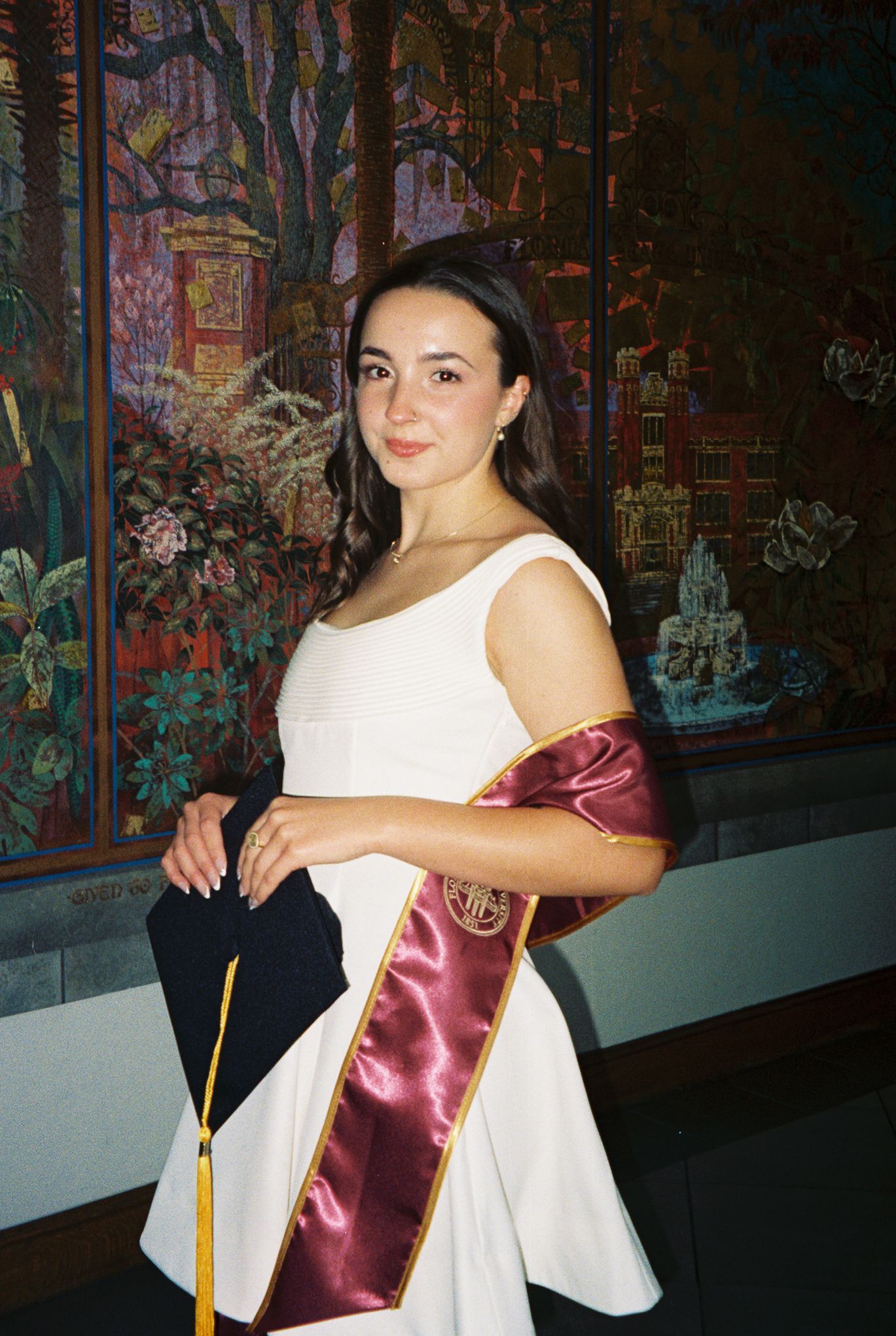 Young woman in white dress with burgundy graduation sash poses proudly in front of colorful mural.