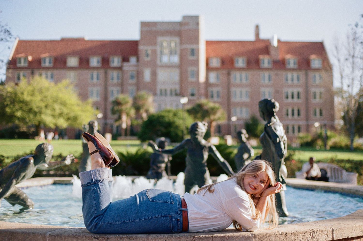 A blonde woman lies on a fountain ledge at a university campus with bronze statues and a red brick building behind her.