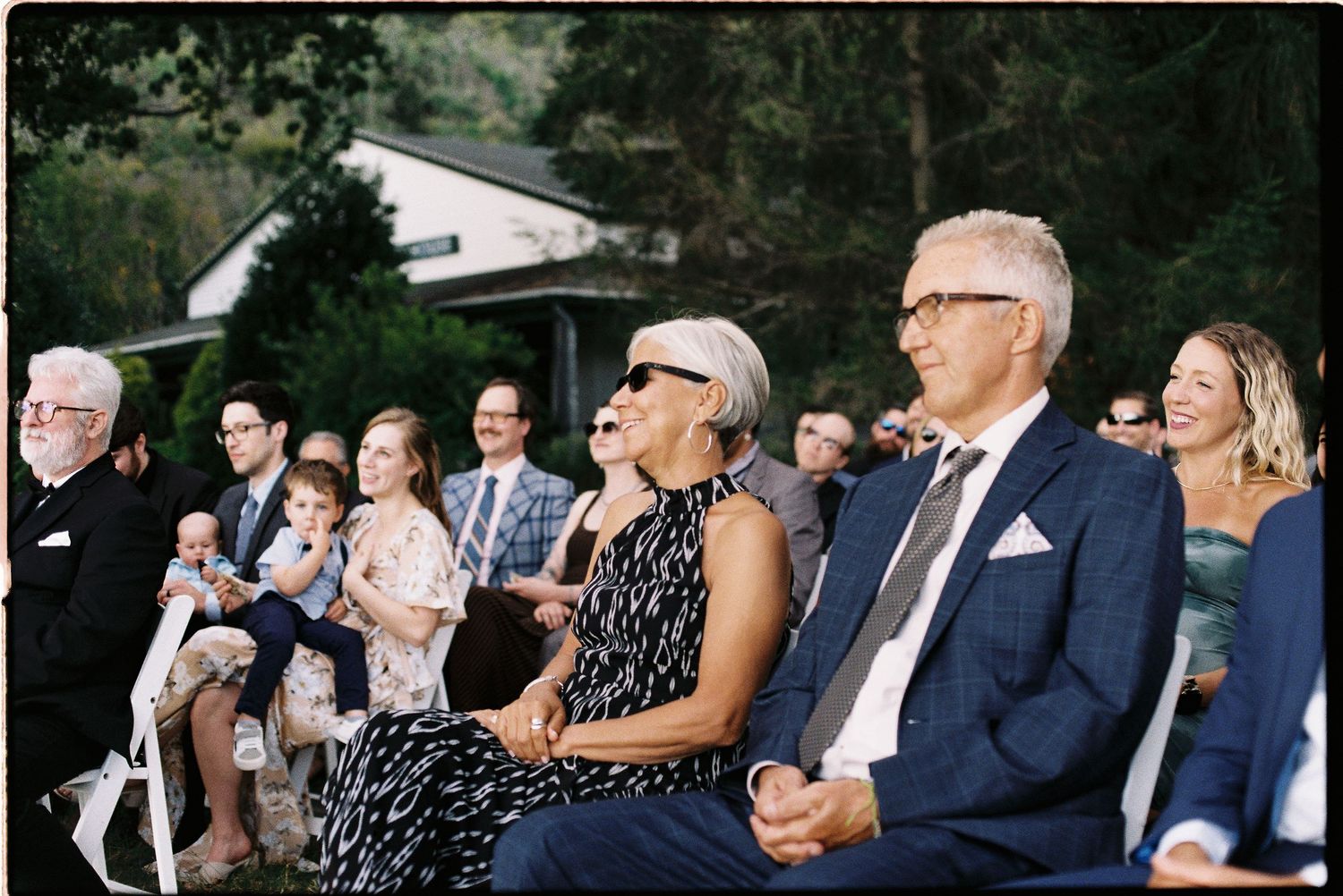 Guests smiling and laughing during an outdoor wedding ceremony seated on white chairs surrounded by lush greenery.