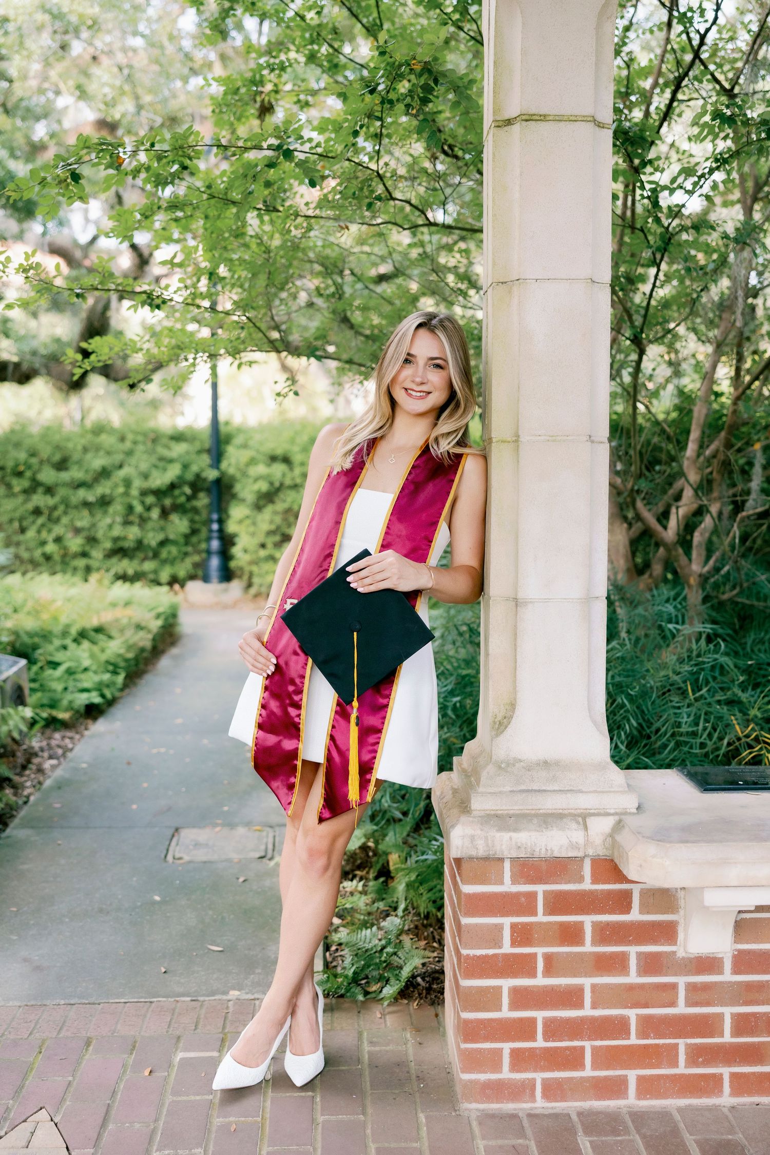 Graduate in red and white dress stands by brick column on campus pathway surrounded by spring blossoms.