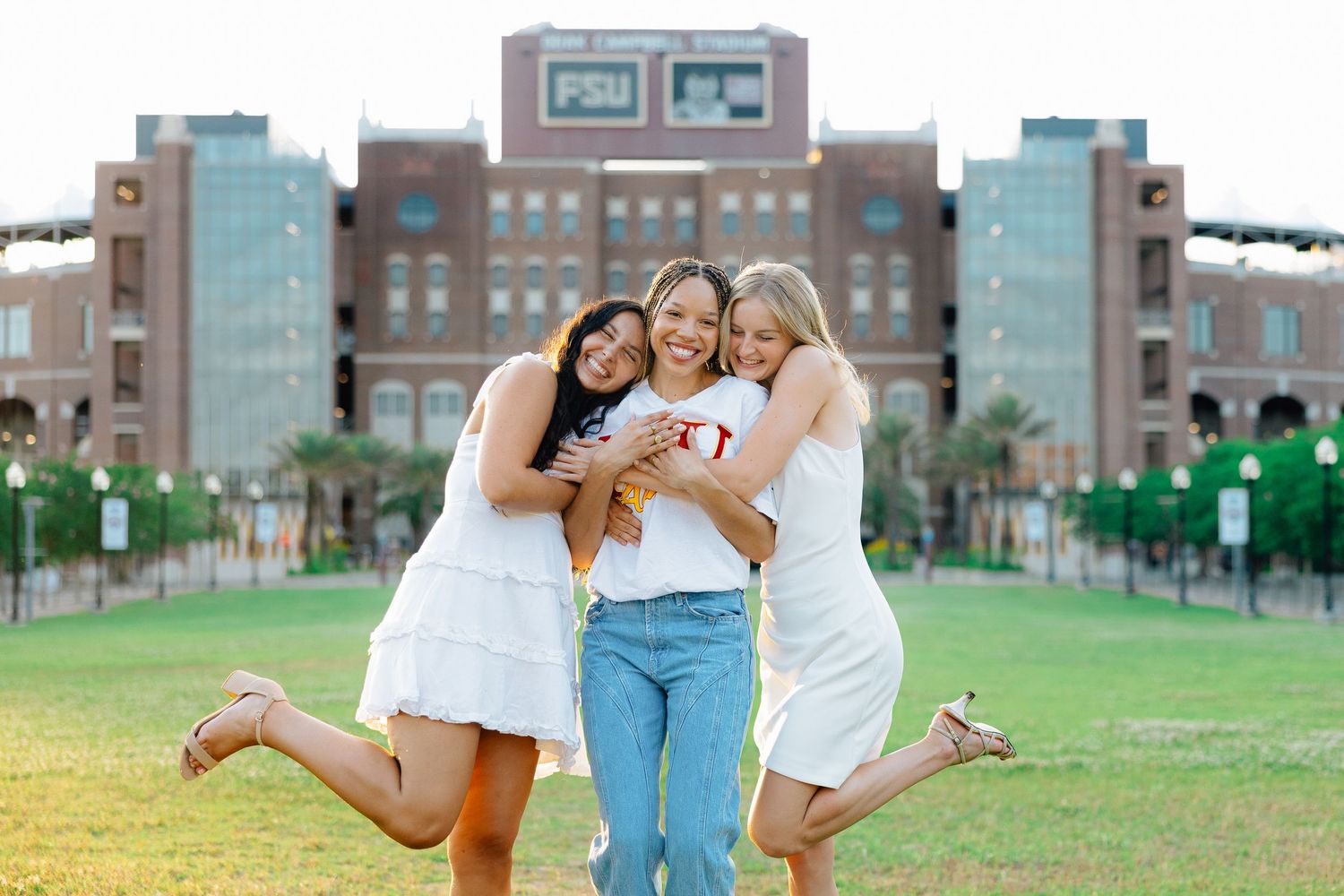 Three friends in casual outfits share joyful moments together on a grassy lawn with brick buildings in the background.