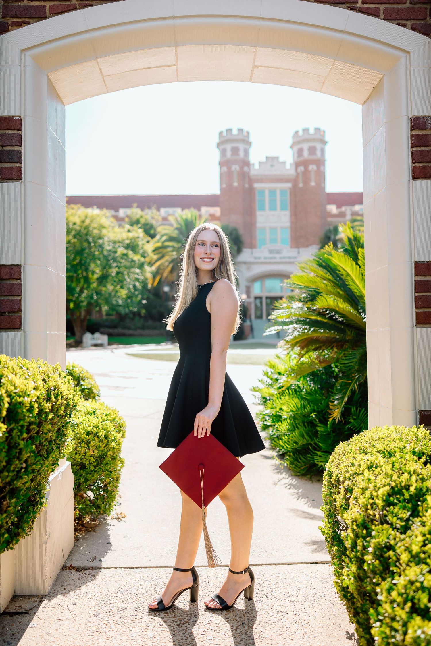 A person in a black top and burgundy skirt poses in front of a white archway and brick castle-like building.