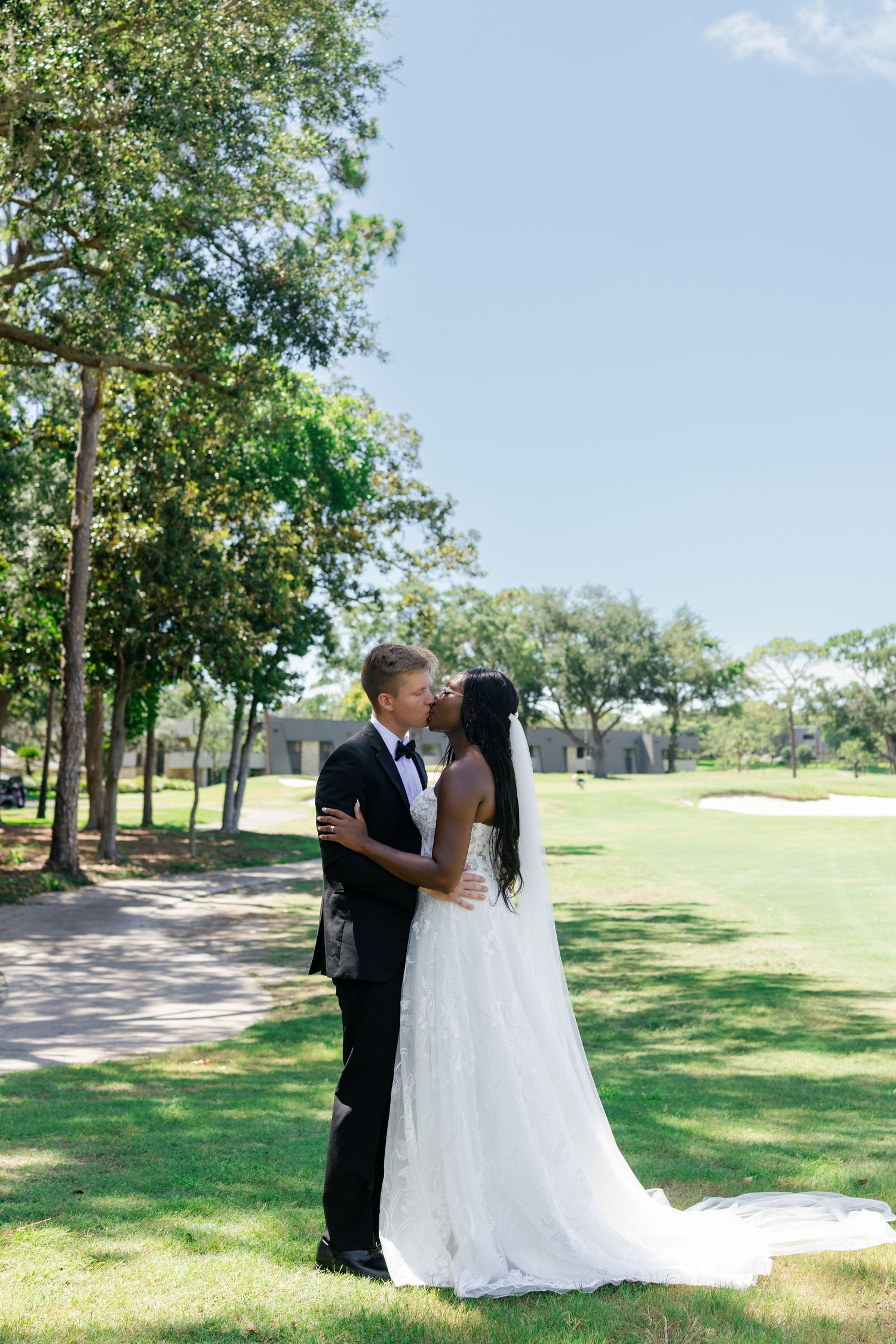 Interracial couple shares romantic kiss on golf course, bride in lace gown with veil, groom in tuxedo.