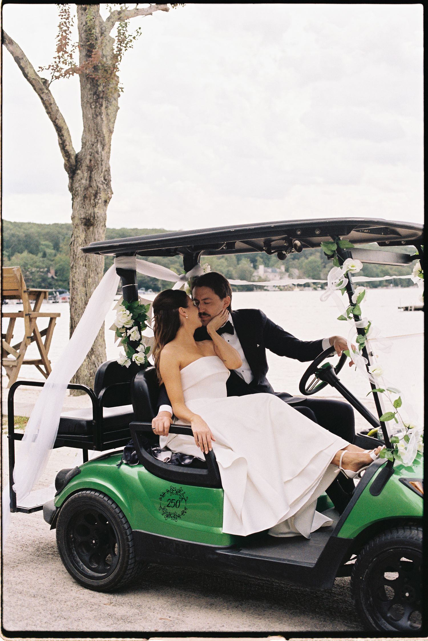 Bride and groom share a kiss on a decorated green golf cart by a waterfront on their wedding day.