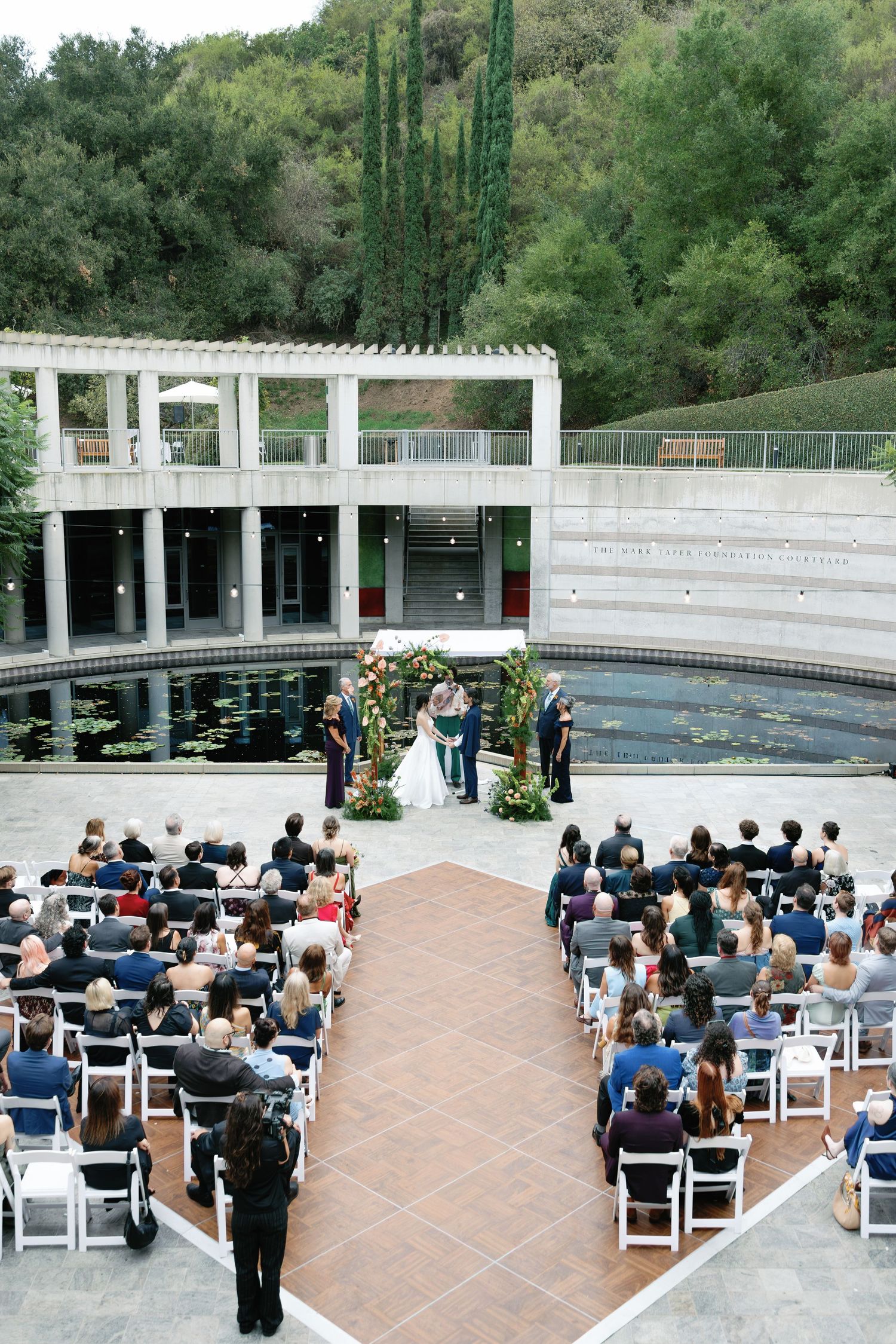 Outdoor wedding ceremony with guests seated on white chairs around a terracotta aisle, facing a couple beside a reflecting pool and hillside