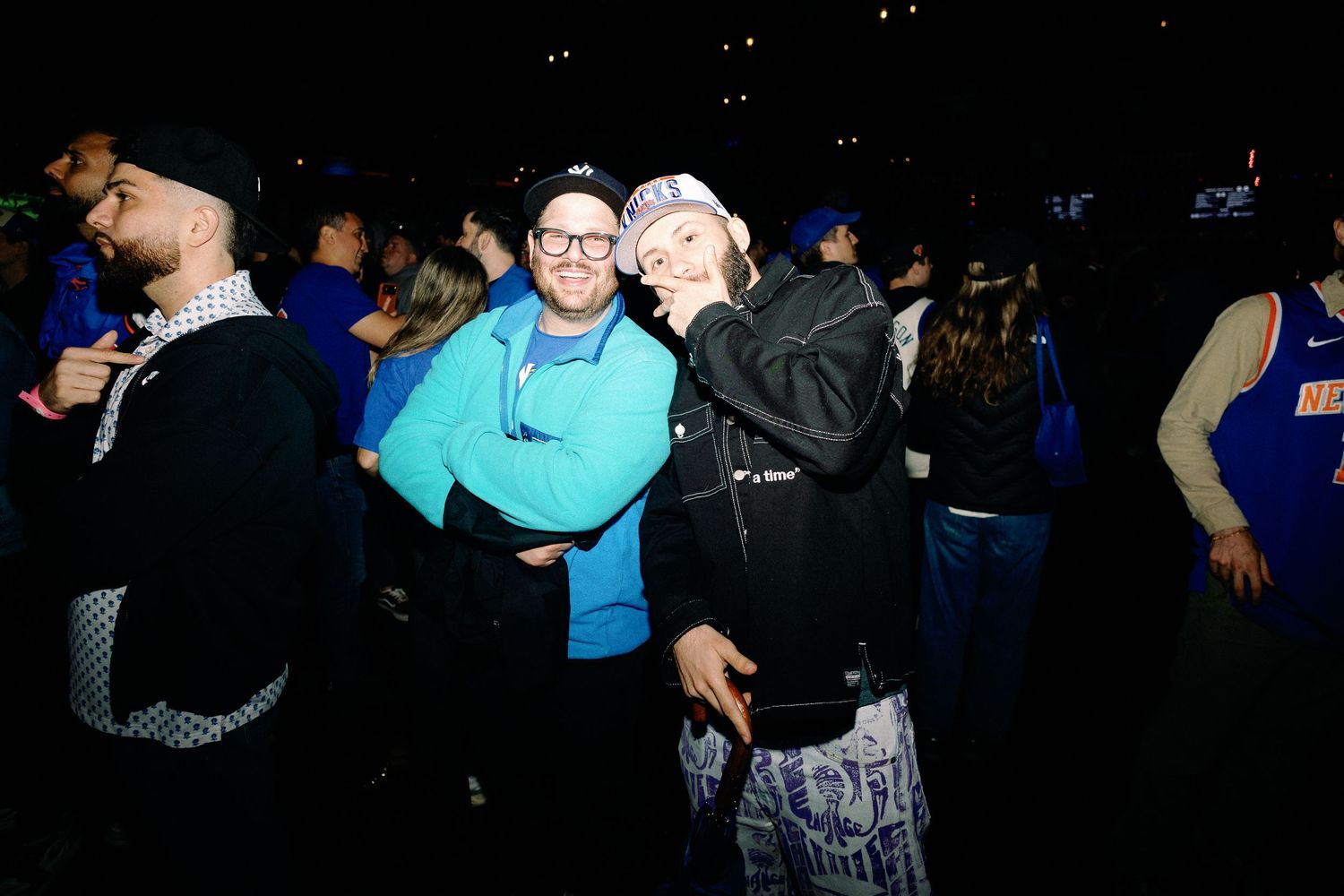 Two friends embrace and smile at a lively nighttime outdoor event with a crowd in blue attire.