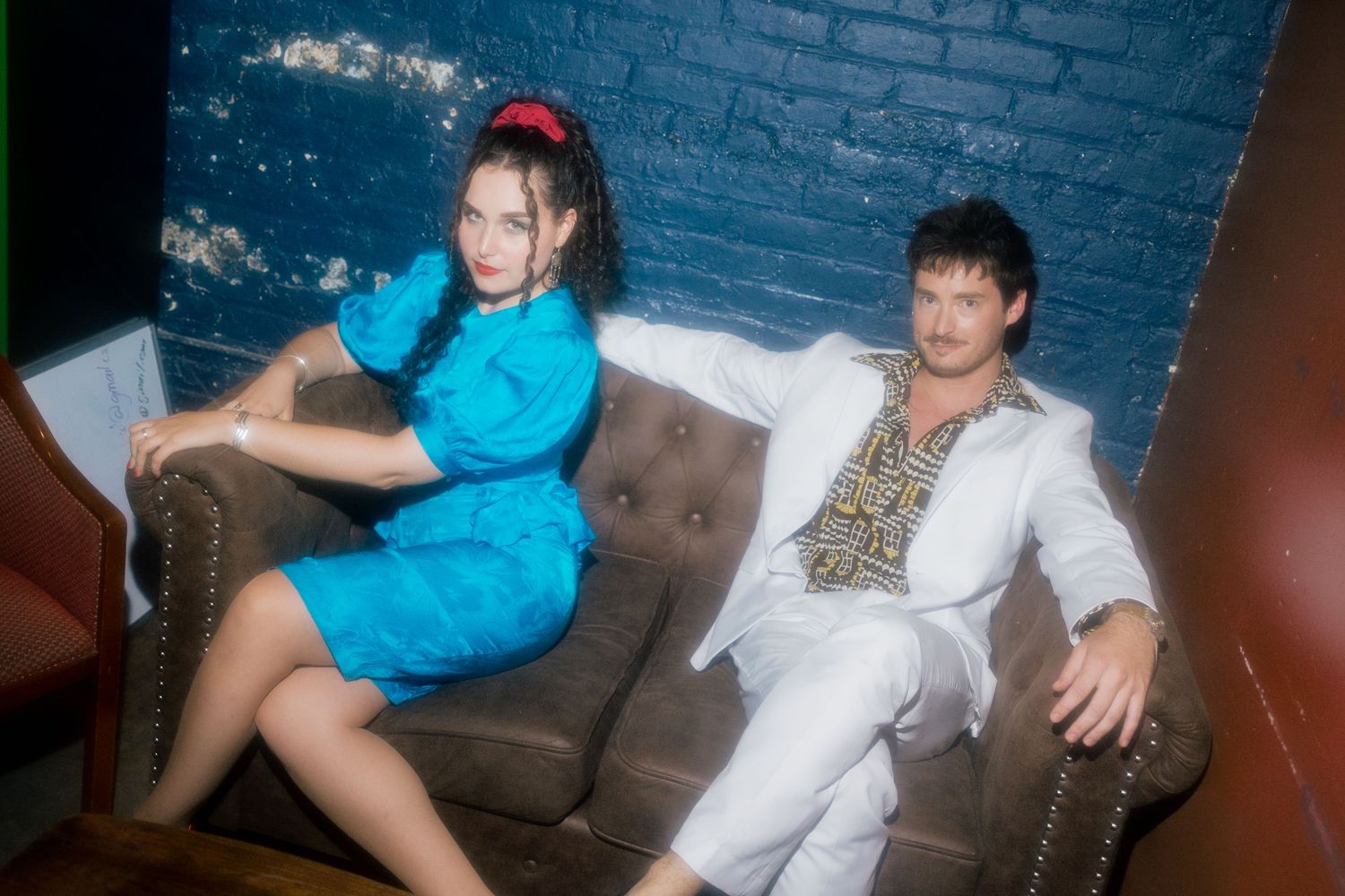 A woman in a blue dress and a man in a white suit pose on a leather couch in a dimly lit room.
