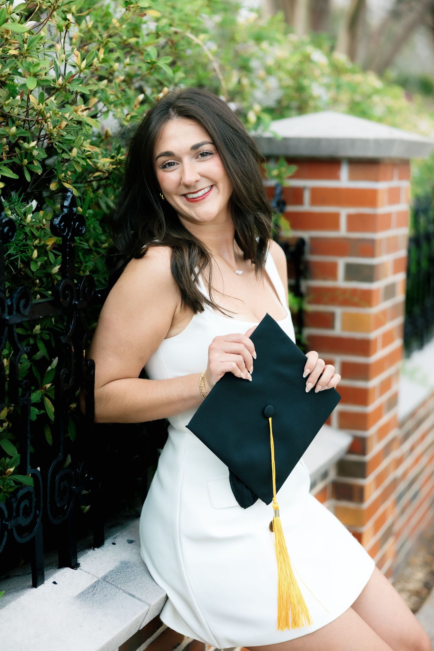 Smiling female graduate in white dress holding black cap with gold tassel outdoors near brick wall and greenery.