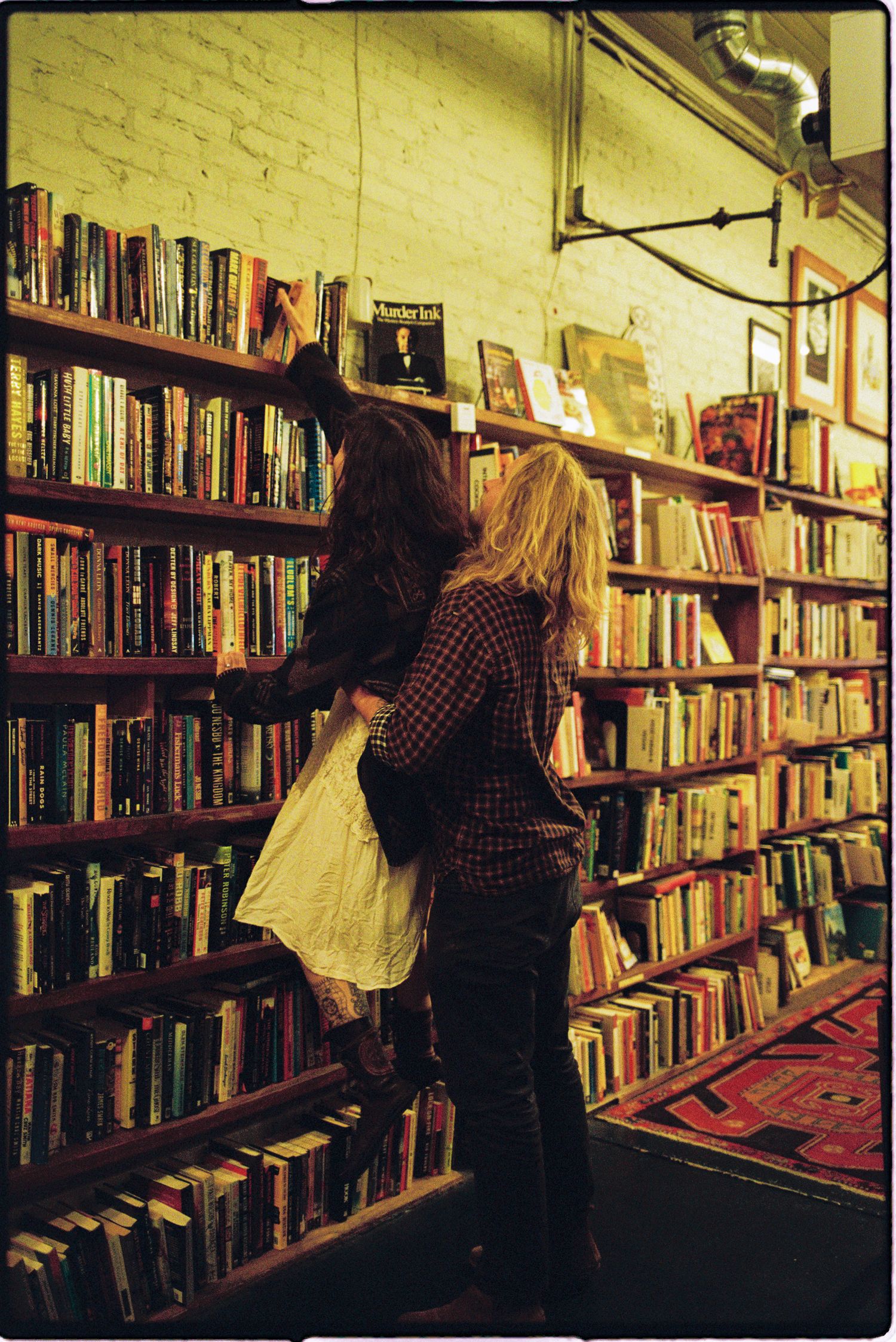 Two women browse crowded shelves of books in a cozy vintage bookstore with brick walls and a red patterned rug.