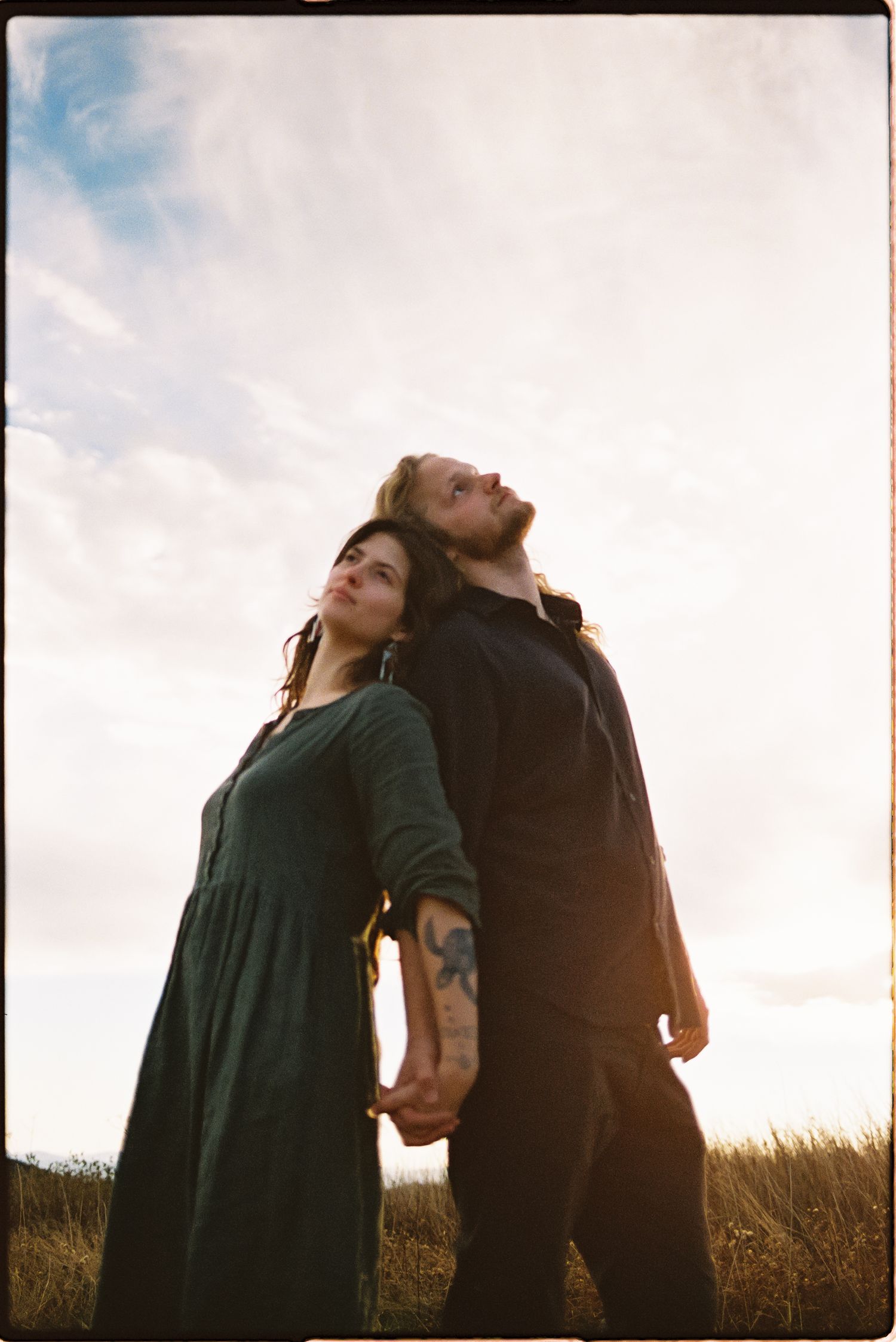 Couple standing back to back holding hands in a grassy field, gazing upward under a soft cloudy sky in this dreamy film-style portrait.