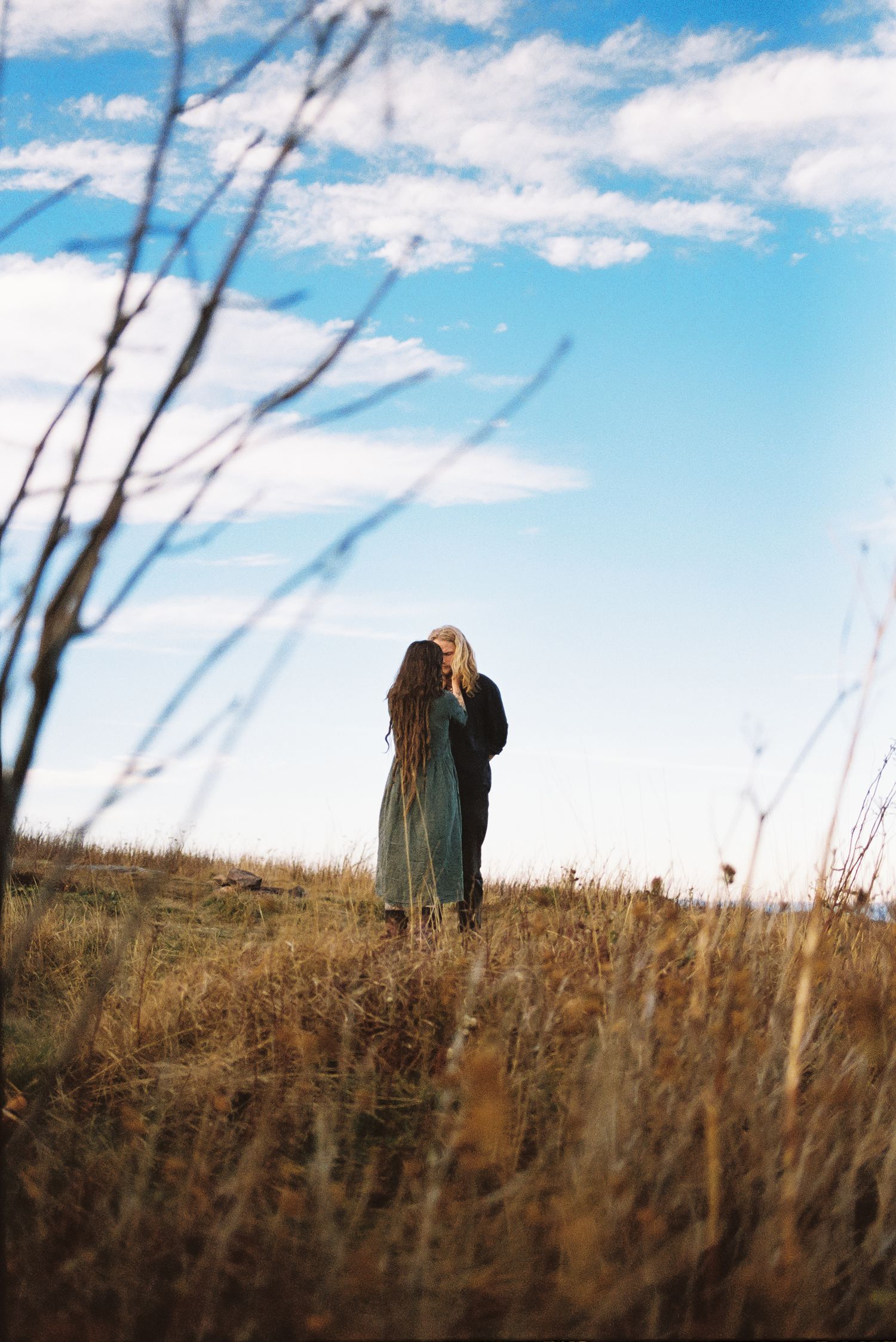 Two people with long hair stand together in a golden grassy field beneath a bright blue sky with wispy clouds, framed by bare branches.