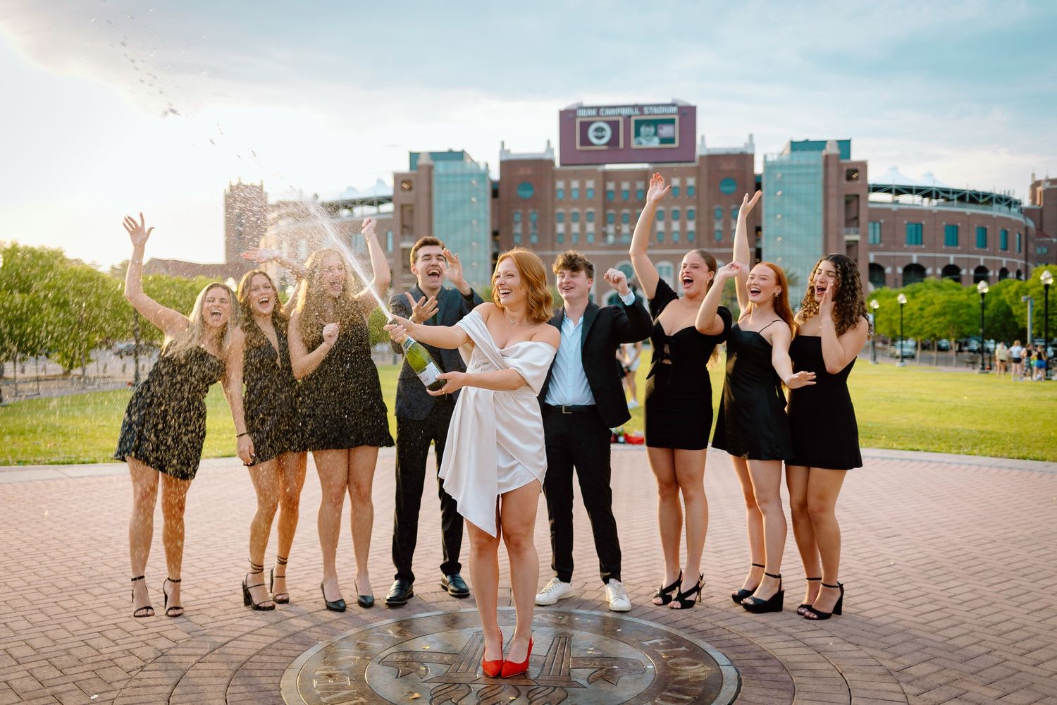 A celebratory group in formal attire poses together and cheers in front of a large building on a sunny day.