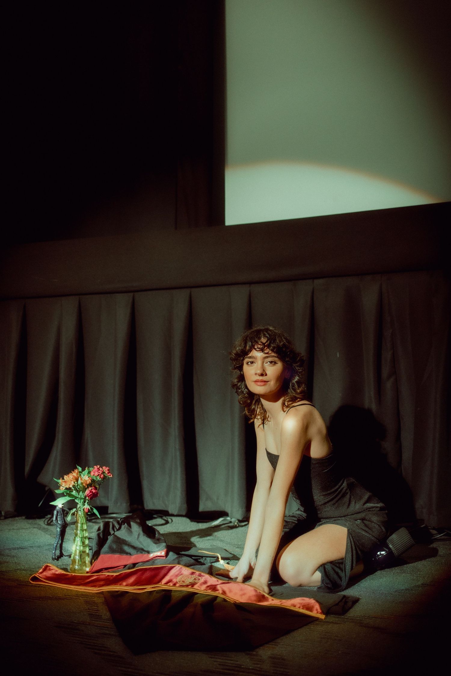 Artistic portrait in moody lighting with figure posing on floor next to vase of flowers on red fabric.