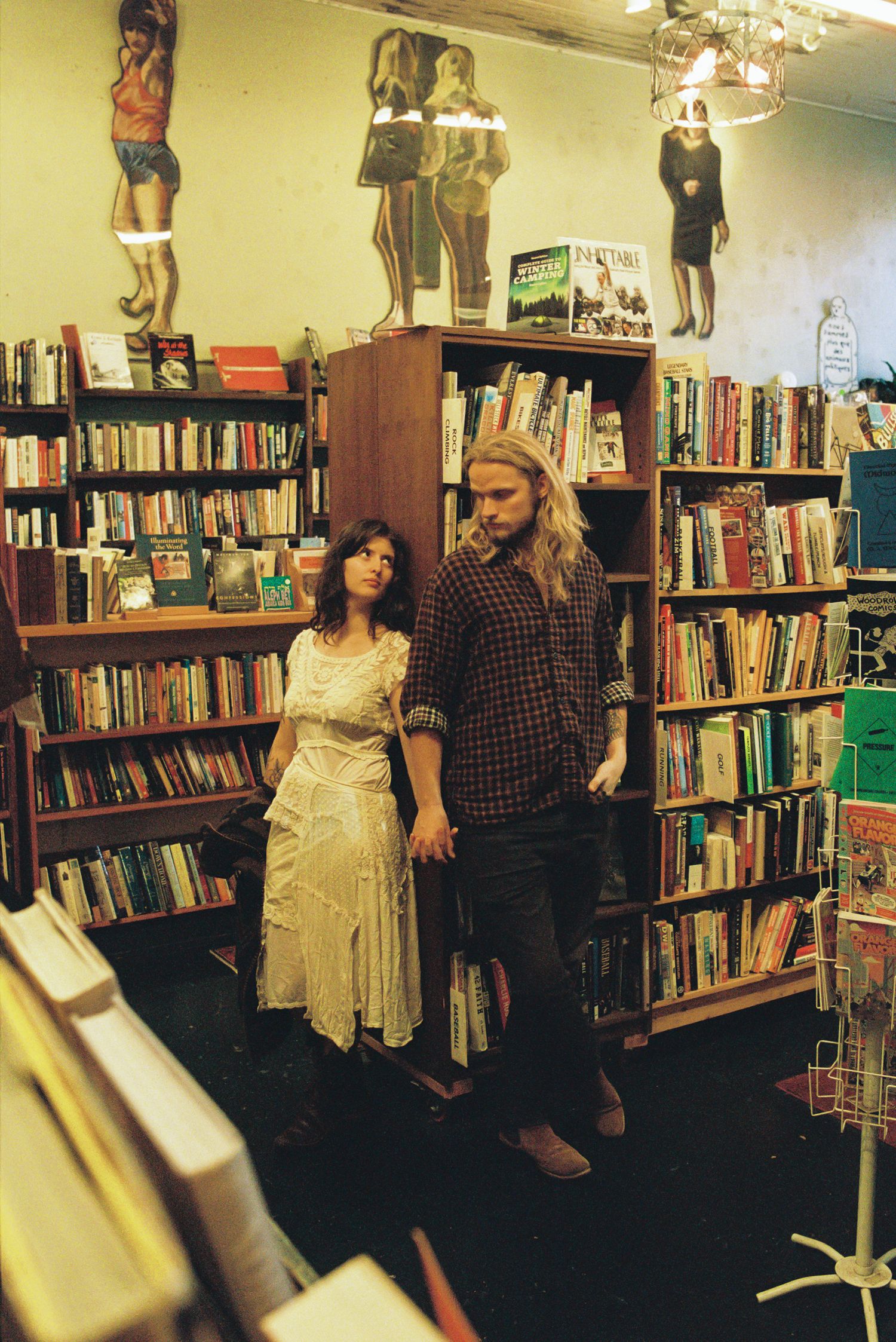 A woman in a cream dress and a man with long hair in a plaid shirt hold hands in a cozy bookstore lined with wooden shelves of books.