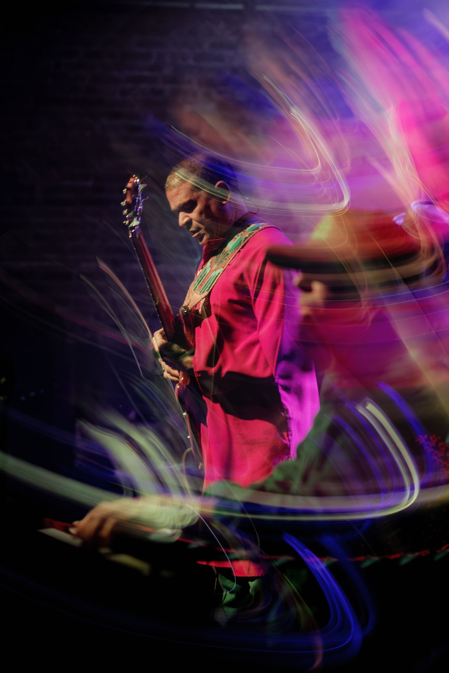 Guitarist in pink jacket performs on stage with vibrant light trails swirling around in a dynamic long-exposure concert photo.