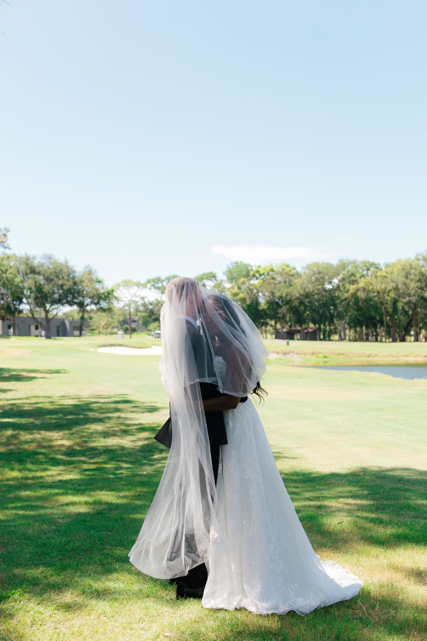 Bride and groom share a kiss under a flowing veil on a golf course lawn on their wedding day.