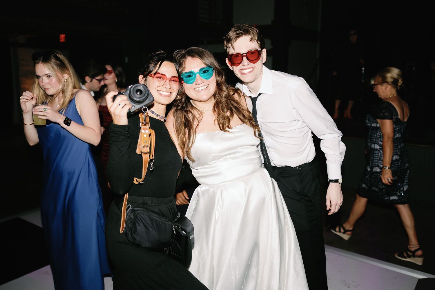 Three friends wearing colorful heart-shaped sunglasses pose together at a wedding reception dance floor.