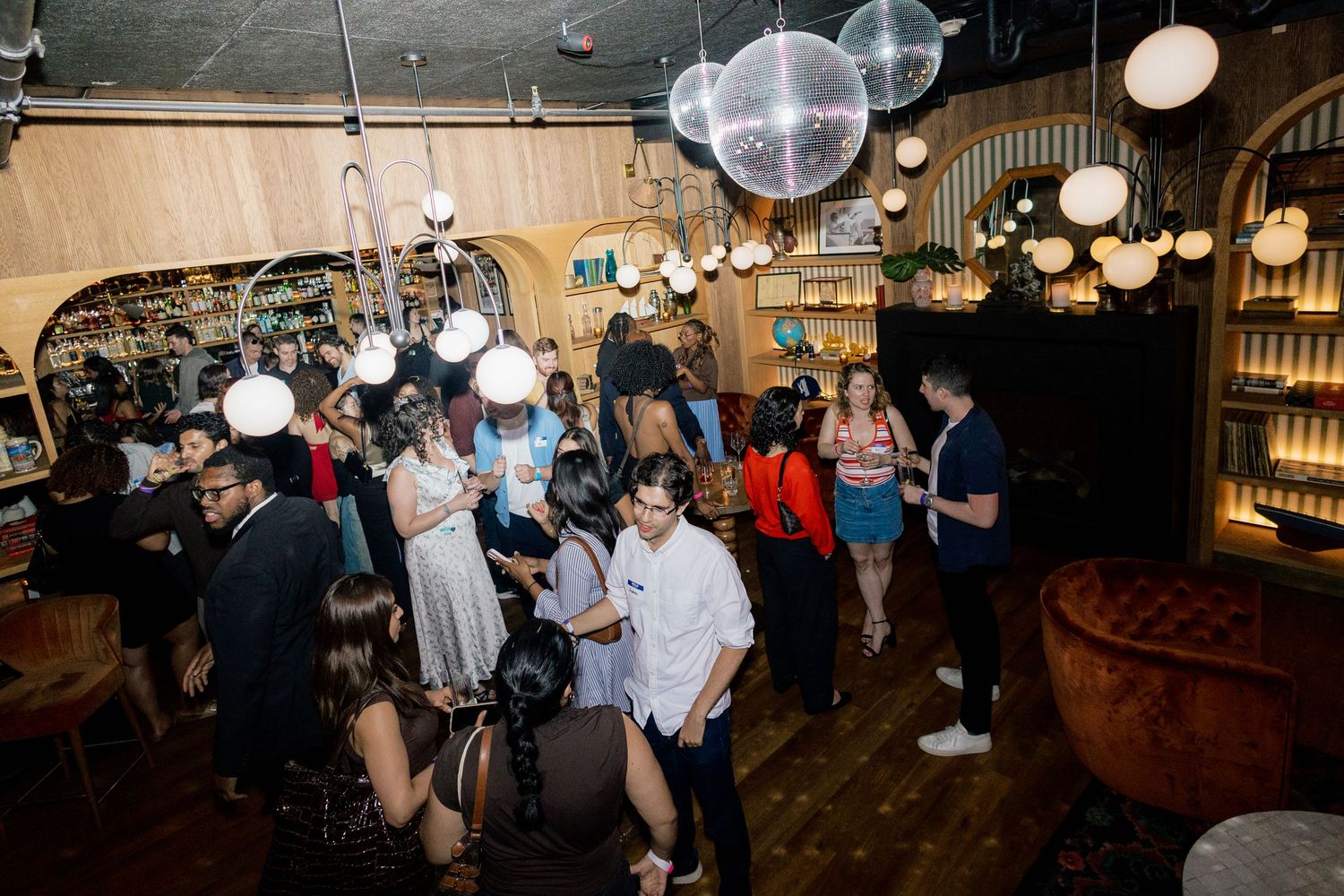 Guests mingle at a trendy bar venue with disco balls, globe lights, and warm wood decor during a lively evening event.