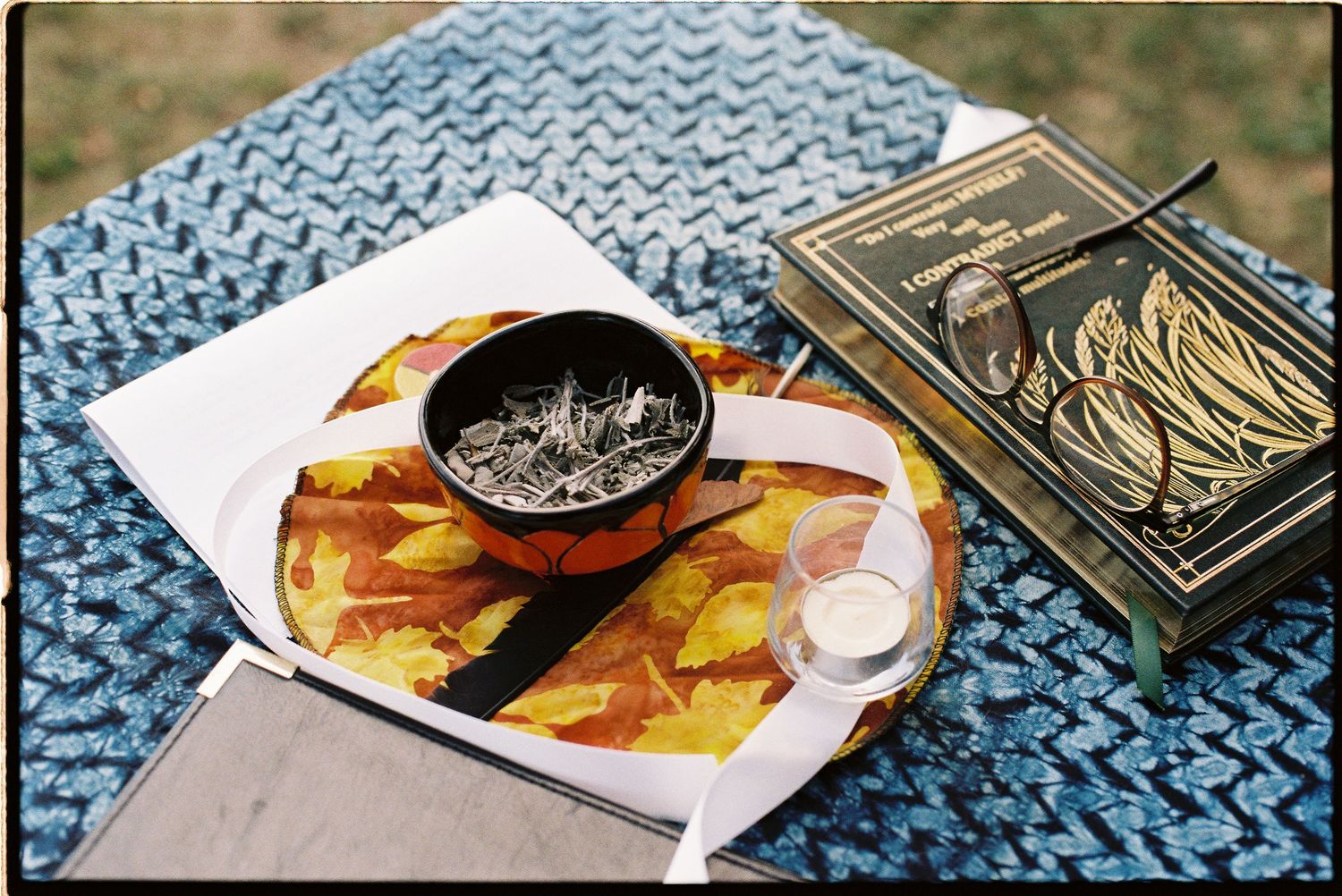 A decorative tray with a bowl of herbs, autumn leaves, and a candle beside an ornate gold book on blue fabric.