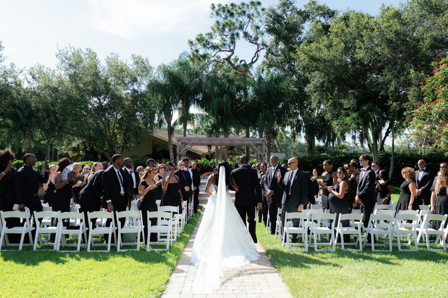 Outdoor wedding ceremony on green lawn with white chairs, guests in formal attire, bride in white gown walking down aisle.