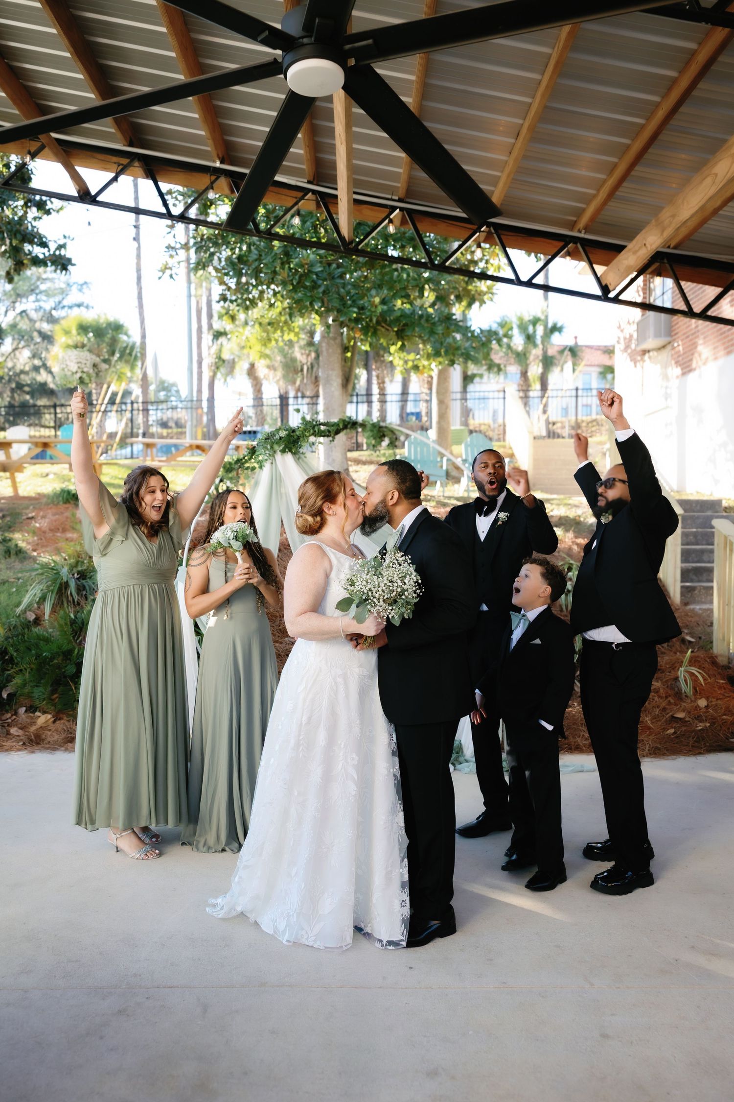 Wedding party laughs together during outdoor photo session with bridesmaids in sage green dresses and groomsmen in black suits.