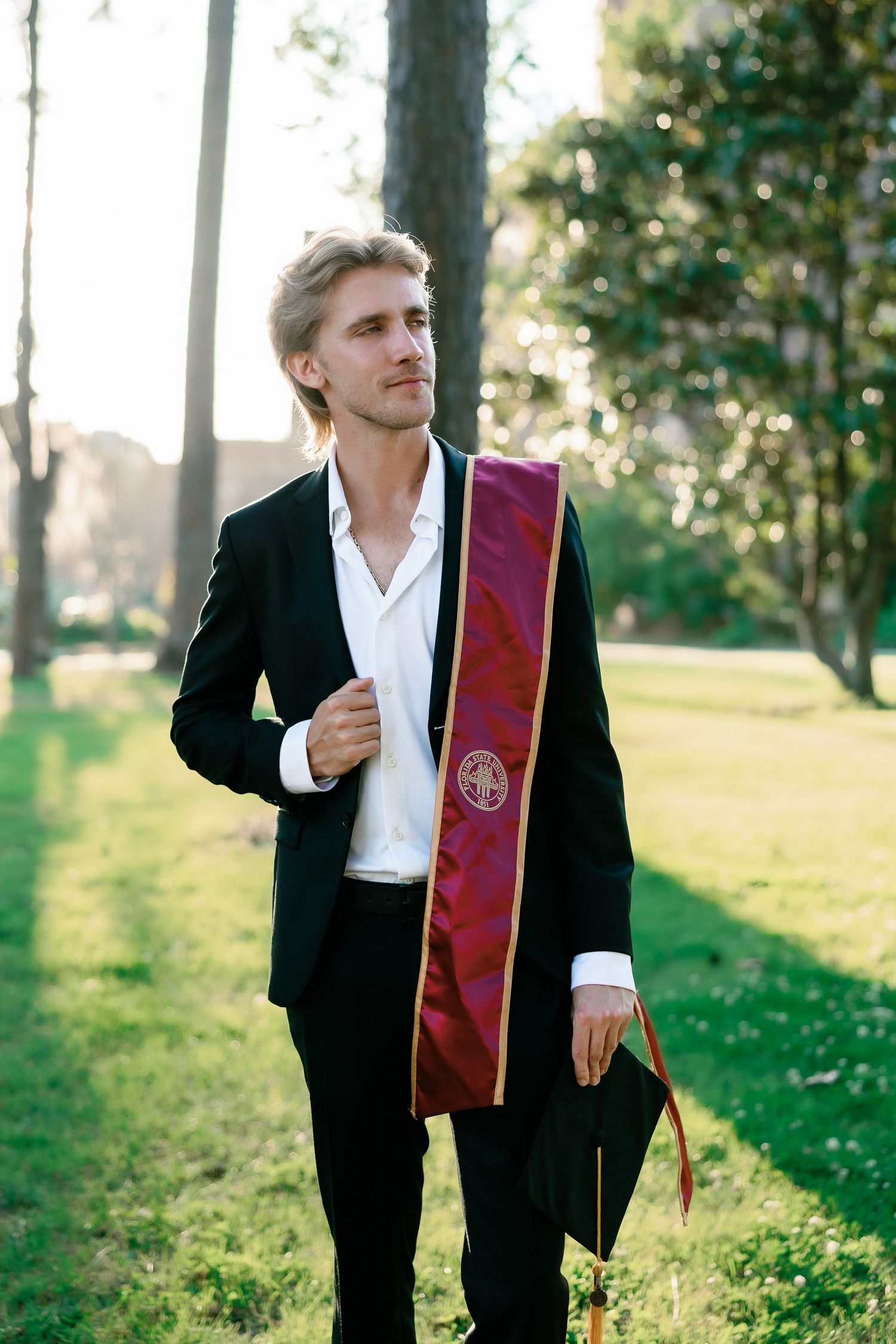 Young man in black suit with maroon graduation stole holding cap outdoors in sunlit park setting.