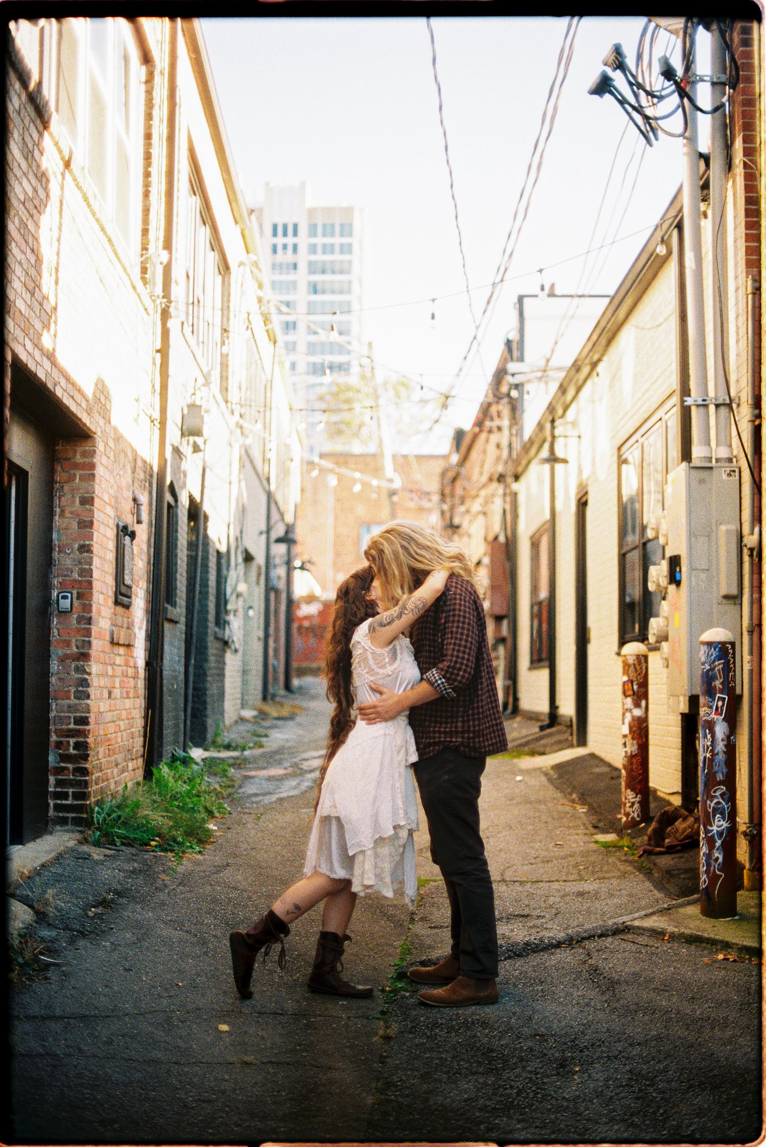 Couple in a white dress and plaid shirt embrace in a brick alley with string lights and a city skyline behind them.
