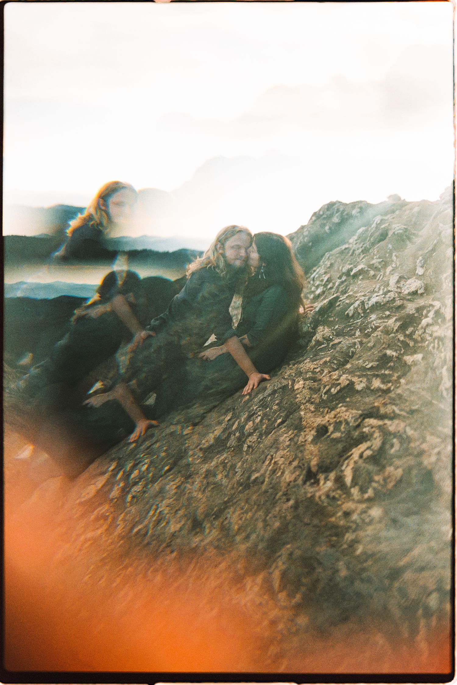 Two people with long hair embrace on a rocky coastal outcrop in a dreamy double-exposure film photo with warm orange light leaks.