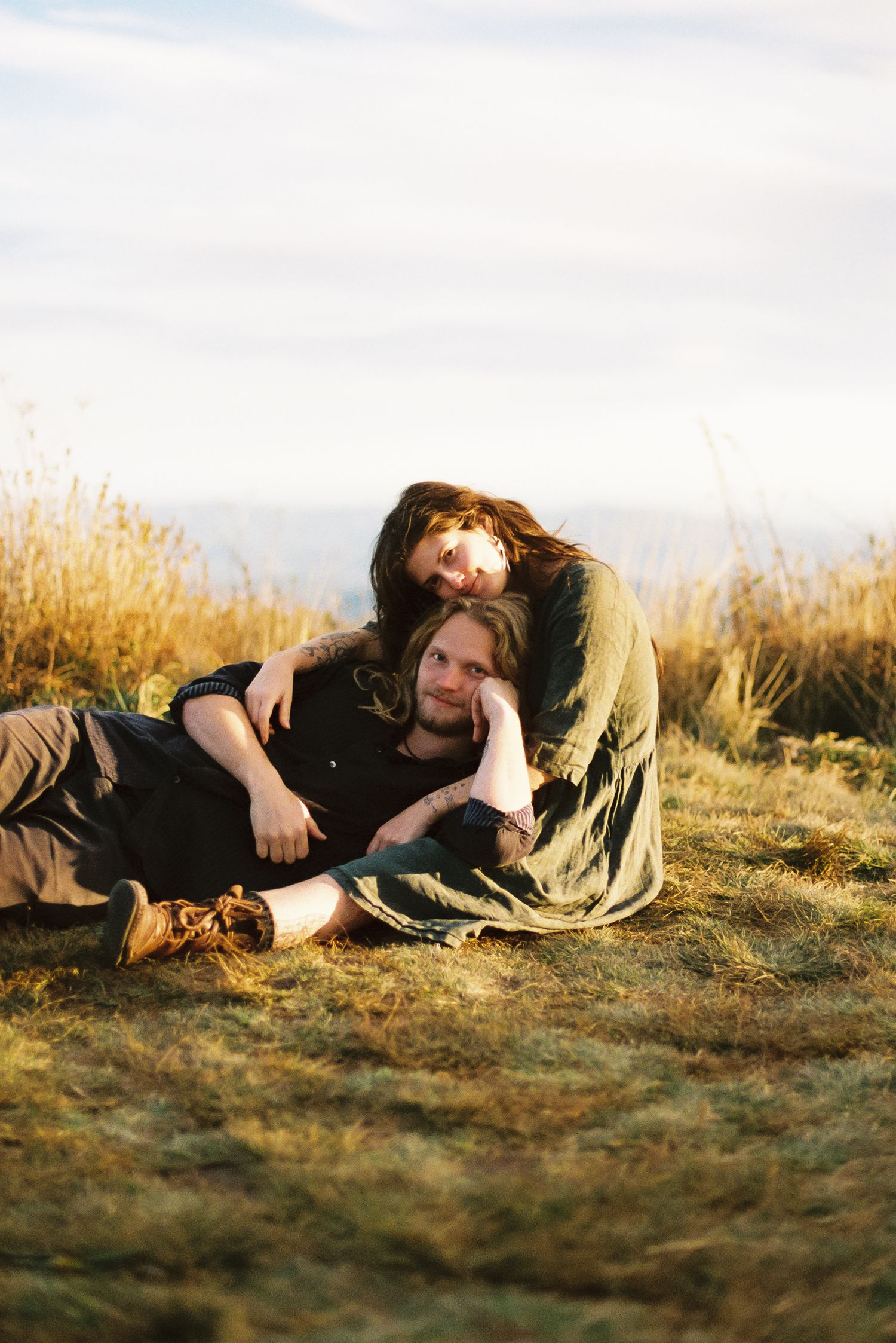 Couple in dark clothing relaxing together on a grassy hillside at golden hour, with tall dry grass and a soft cloudy sky behind them.