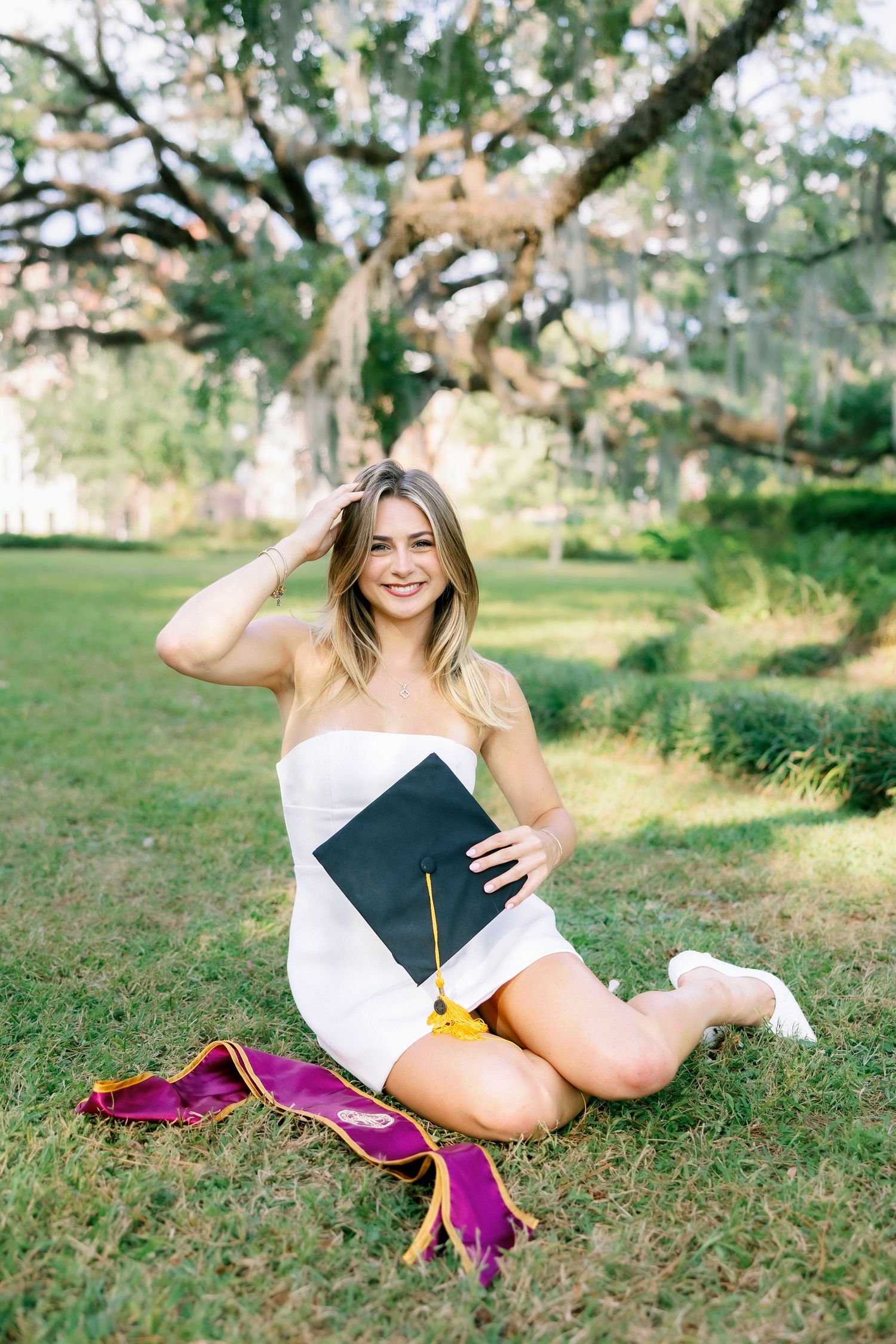 Graduate in white dress lays in grass with diploma and magenta graduation stole during outdoor photo session.