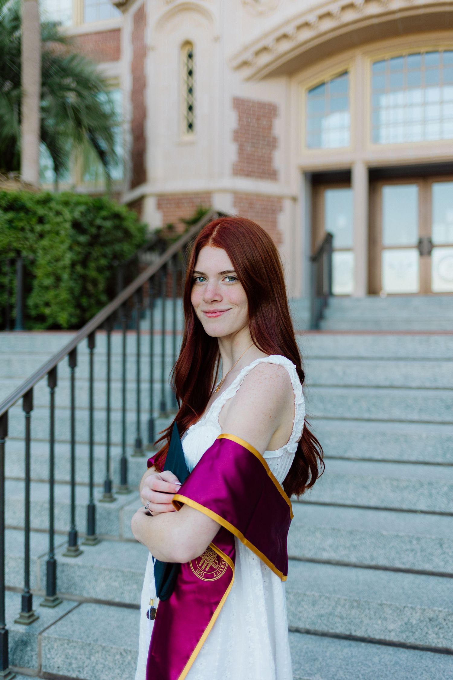 Graduate in white dress and purple graduation stole poses on stone steps in front of building with palm trees.