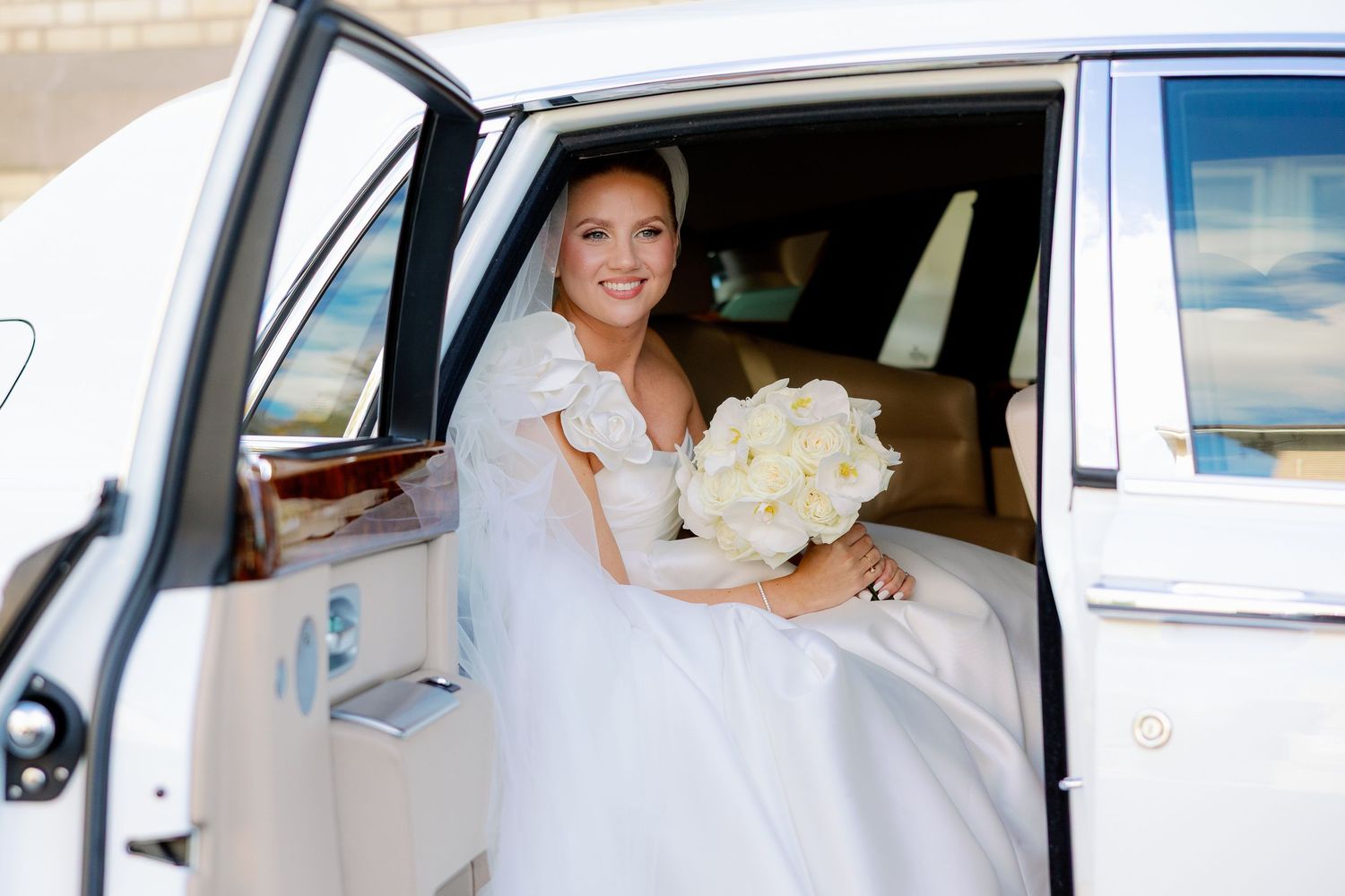 A bride in a white wedding dress sits in the back seat of a car holding a bouquet of white flowers.