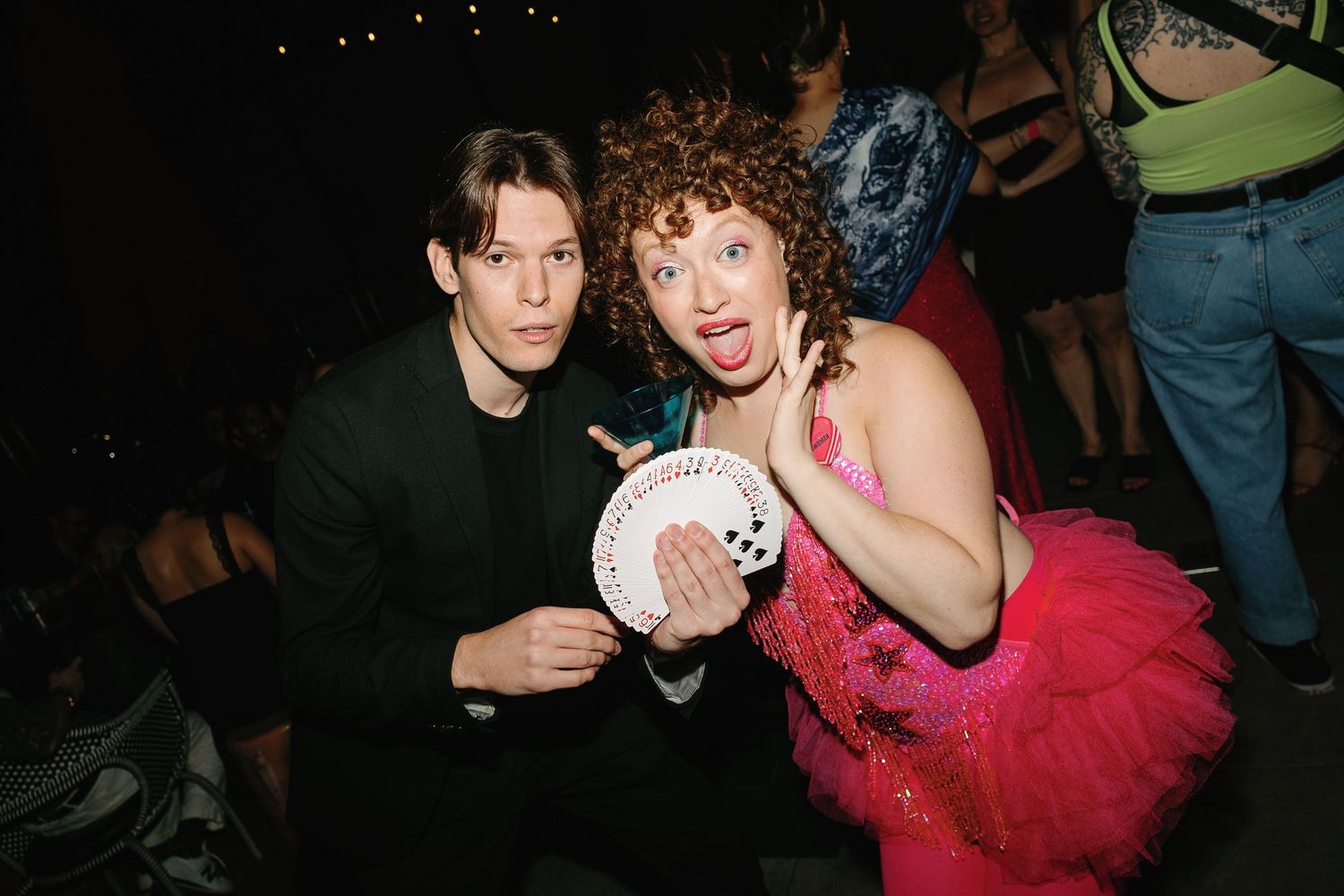 Two people in costumes pose at a party, one in a black suit and one in a pink tutu holding playing cards.
