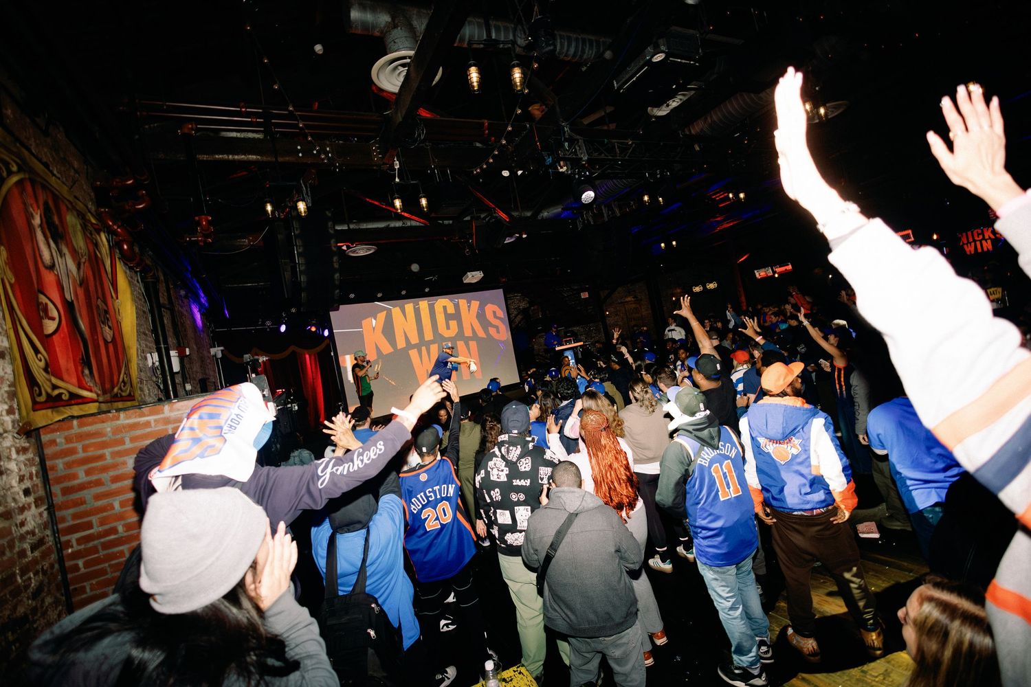 Excited New York Knicks fans celebrate together in a packed bar, raising hands with a Knicks sign visible.