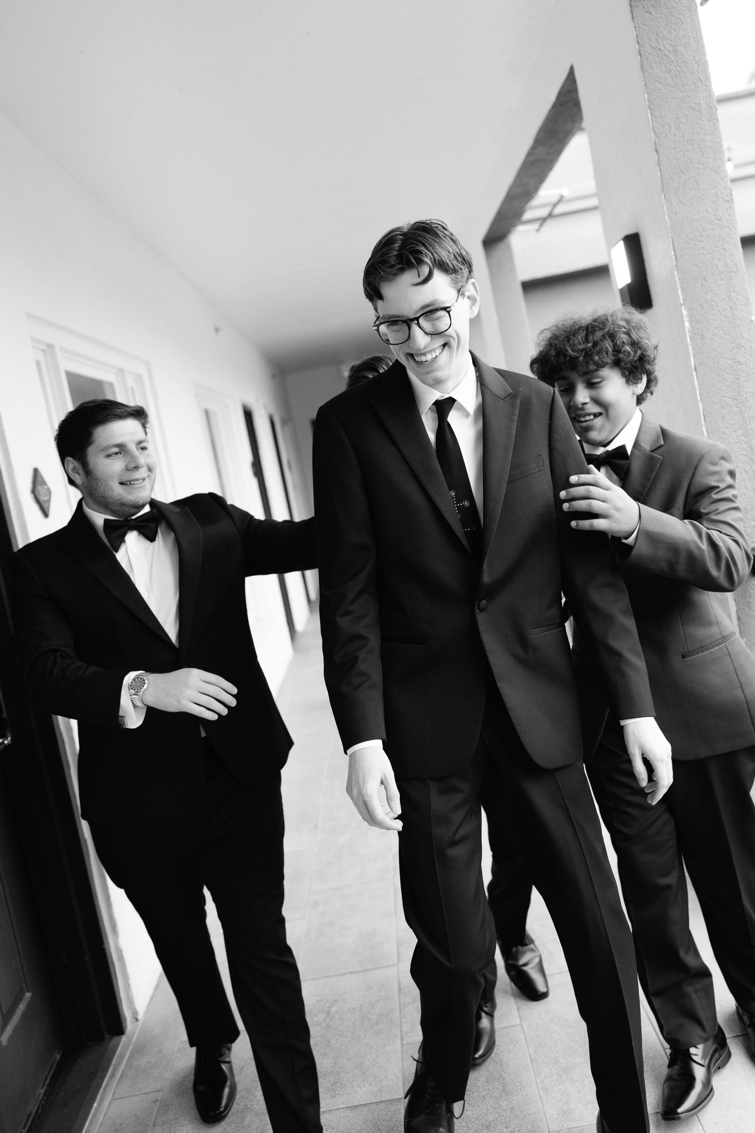 Three young men in formal suits and bow ties laughing together in a hallway, black and white photo.