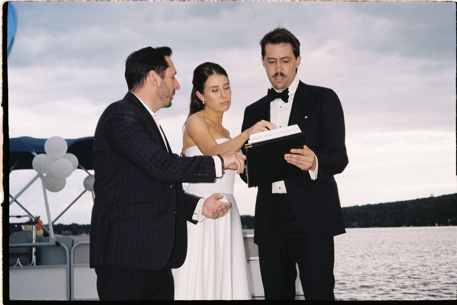 Couple exchanging wedding vows on a boat with an officiant holding a book, water and cloudy sky in background.
