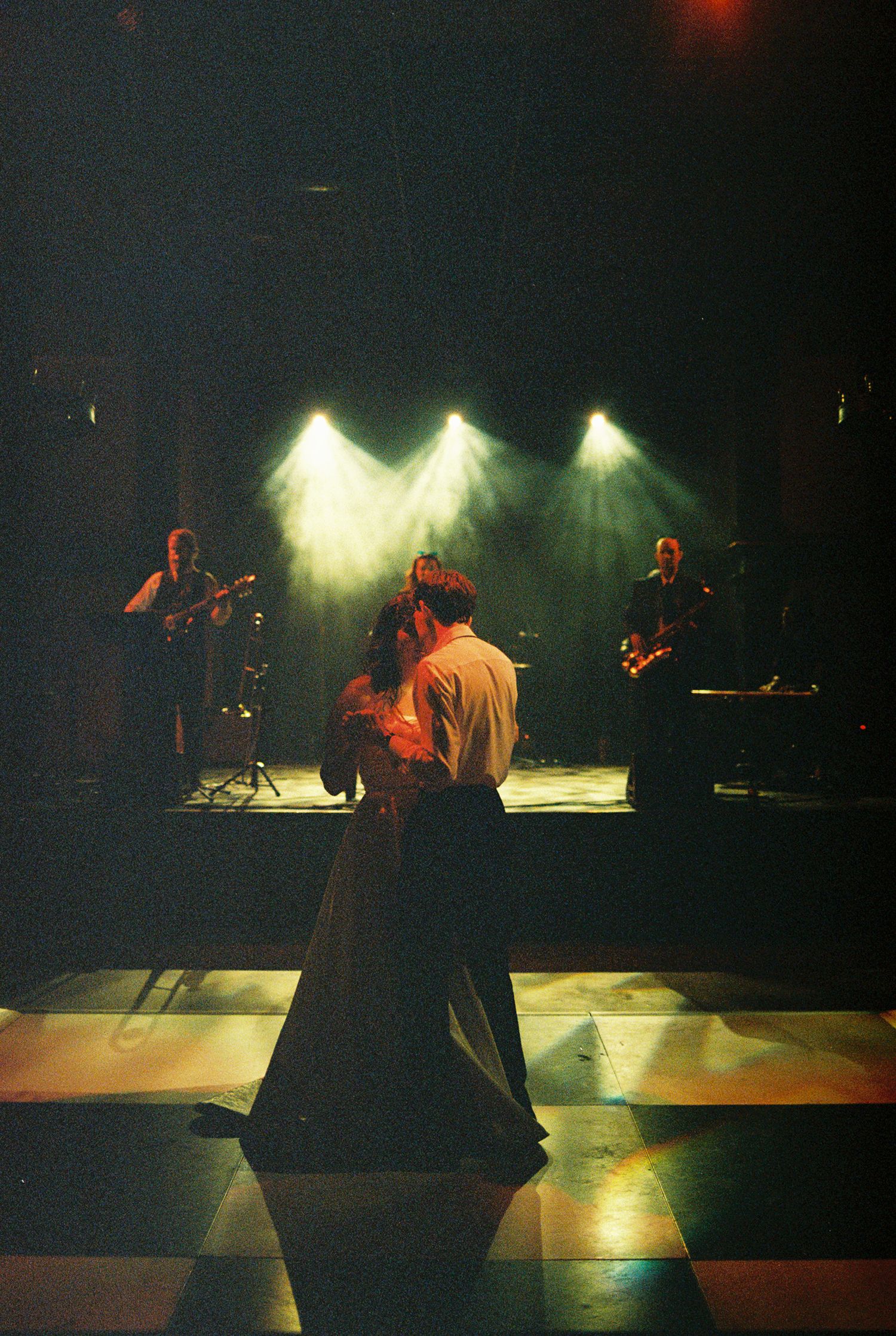 Couple slow dancing on checkered floor under spotlights with live band performing on stage in dimly lit venue.